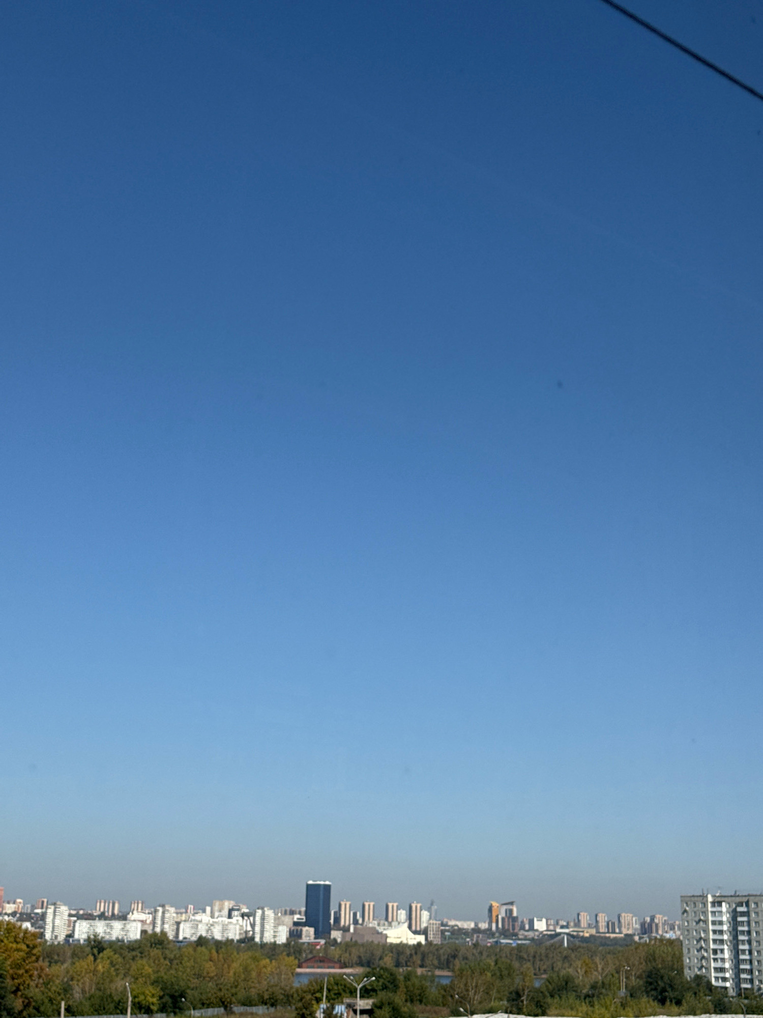 A wide, clear blue sky dominates the upper two-thirds of the image. The lower third depicts a city skyline comprised of numerous apartment buildings and some taller, modern structures, interspersed with trees and some greenery. A dark, thin cable runs diagonally across the top right portion of the sky. The image appears to be taken from an elevated vantage point, looking out over the city, with the buildings in the background suggesting a mid-to-late autumn season due to the colors of the foliage. The scene is tranquil and lacks any visible human presence or activity. The overall impression is one of a daytime urban landscape.