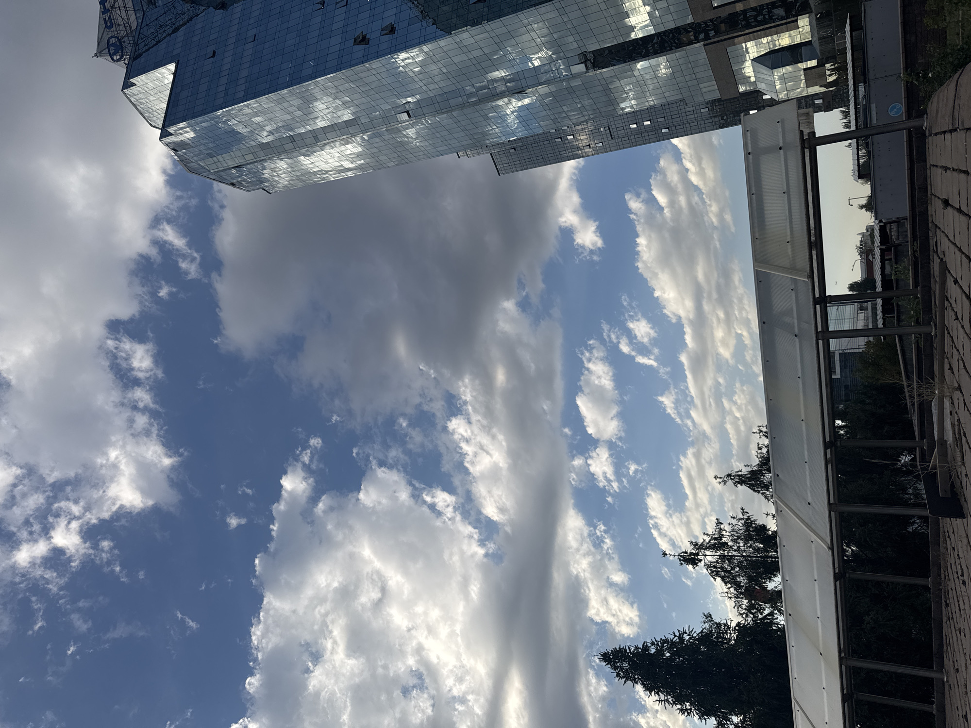 The image captures a street-level view of a city with a prominent glass-fronted skyscraper on the right. The skyscraper, reflecting the sky and clouds, rises high into a blue sky dotted with white and grey cumulus clouds. In the foreground, a covered walkway with a translucent roof is visible, supported by metal poles. Dark green coniferous trees are silhouetted behind the walkway and in the left mid-ground. The ground in the foreground appears to be paved with bricks. In the distance, other buildings and structures can be seen, suggesting an urban environment. The overall scene suggests an outdoor urban setting, possibly in Almaty, Kazakhstan, given the architectural style of the skyscraper.