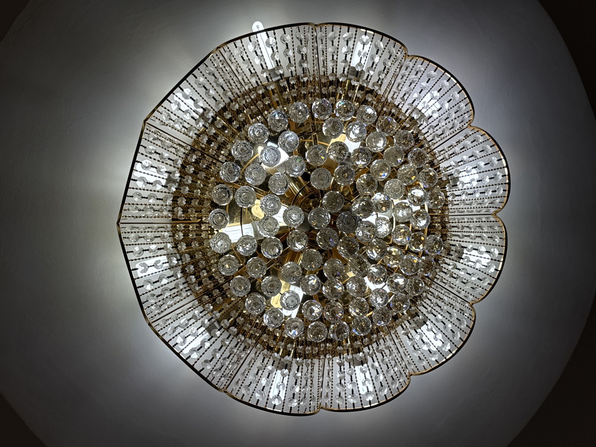A photograph depicts a complex, ornate chandelier mounted on a ceiling. The chandelier features a gold-colored frame with multiple tiers of crystal beads and spherical glass or crystal elements. These elements create a dazzling display of light and reflection. The ceiling is plain and white, providing a contrasting background that accentuates the chandelier's intricate design. The location appears to be indoors within a residential or commercial setting. The city of Unknown, Unknown, could not be determined from the provided image.