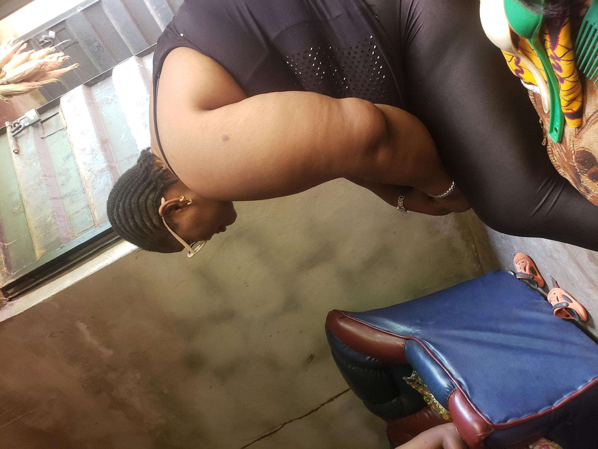 A middle-aged, dark-skinned woman with braided hair is wearing glasses, a black top, and black leggings. She has a silver bracelet on her wrist. She is indoors, near a window with what appears to be corn stalks on the outside ledge. She is seated next to a blue and maroon chair and orange sandals. The location is Umundugba, Nigeria.
