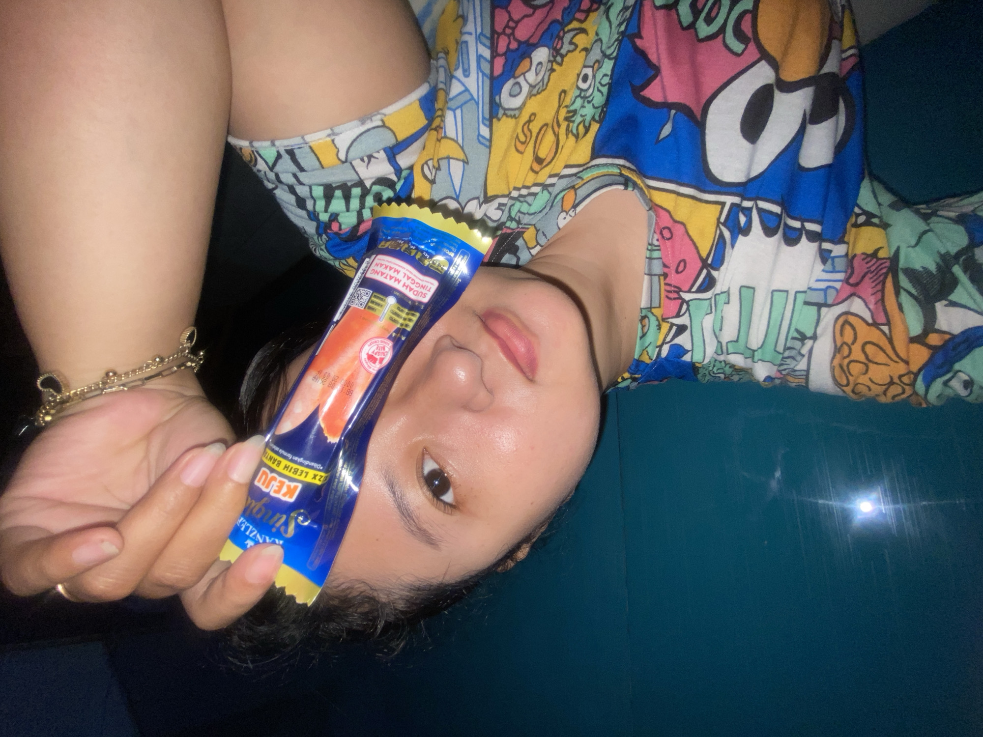 The image depicts a person, appearing to be an adult female, positioned in front of a dark teal wall, likely indoors within the city of Gelam, Indonesia. The individual, visible from the chest up, has light skin, dark hair, and is looking towards the camera with a slight smile. She is wearing a multi-colored t-shirt featuring cartoon-style graphics. Her right hand, with natural nails, holds a blue and yellow Kanzler Single Keju (cheese) sausage packet, which displays an image of a red sausage and text in Indonesian, "SUDAH MATANG TINGGAL MAKAN" (Ready to eat). On her left wrist, several gold-colored bracelets are visible, one adorned with a small black heart charm. A prominent lens flare from a light source is visible on the upper left portion of the teal background.