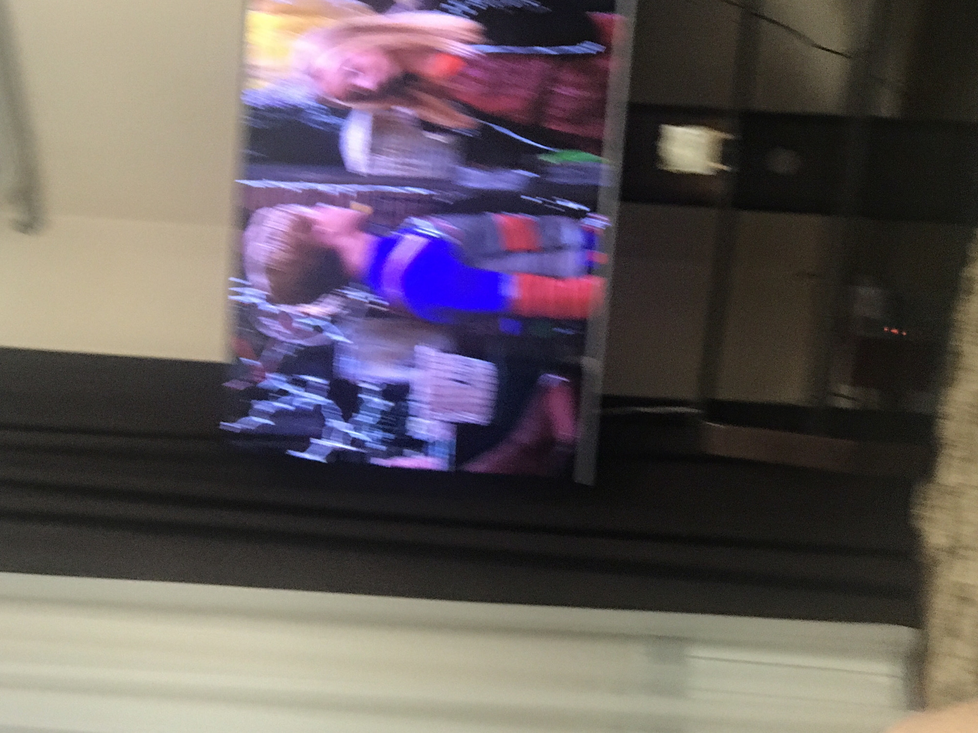 The image shows a blurred television screen displaying two individuals. One person has blond hair and is wearing a red and black top. The other person is wearing a blue and orange top. The television is sitting on a dark-colored surface. A white wall is visible below the dark surface. The environment around the television is indistinct. The location is unknown as the image provides no information about a location in Yerevan, Armenia.