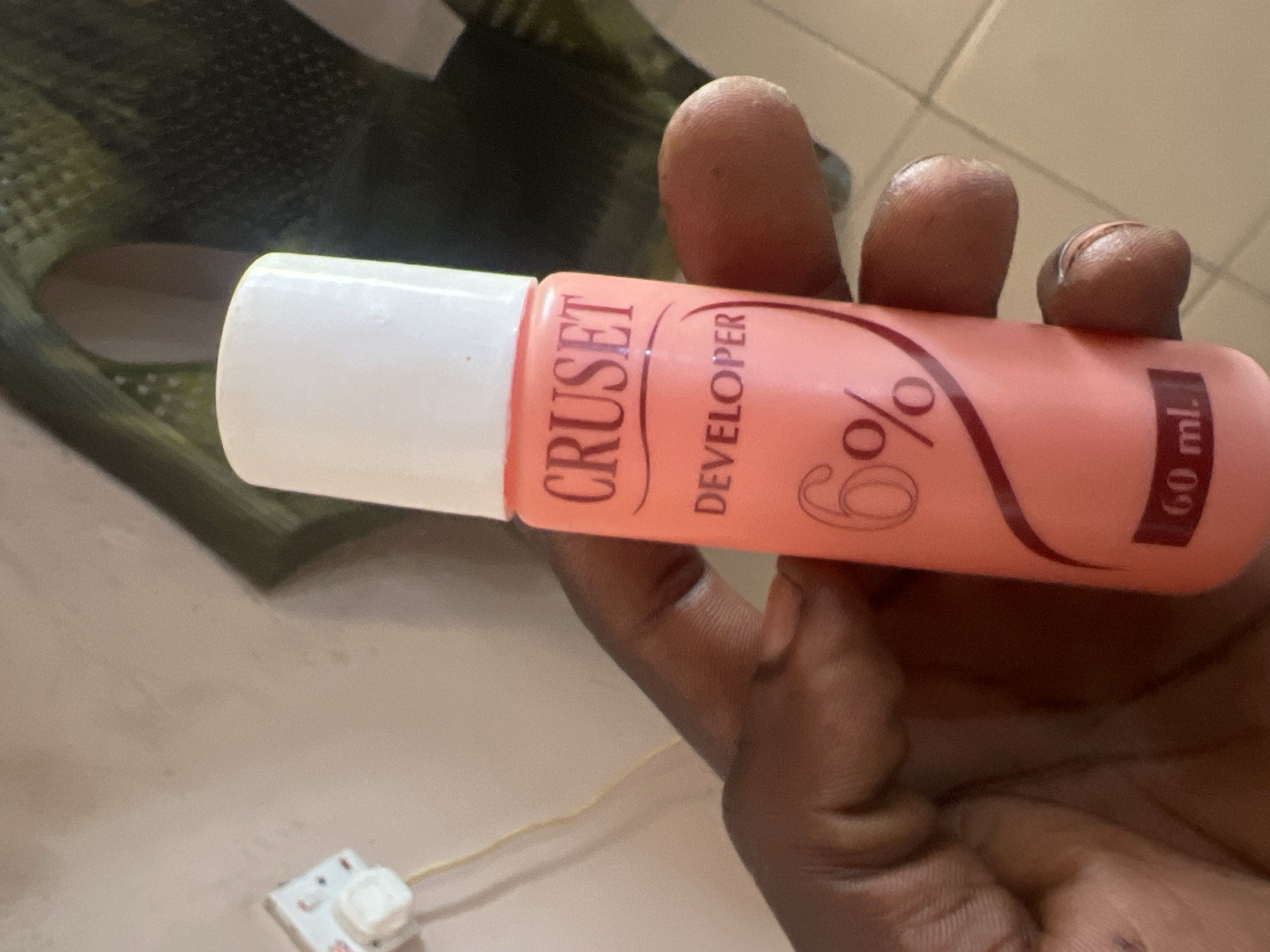 A hand holds a pink bottle of "CRUSET DEVELOPER 6%" with a white cap. The bottle is inscribed with the brand name and product details in dark red lettering. The background is a blurred wall and a green patterned fabric. The location is Jalingo, Nigeria.