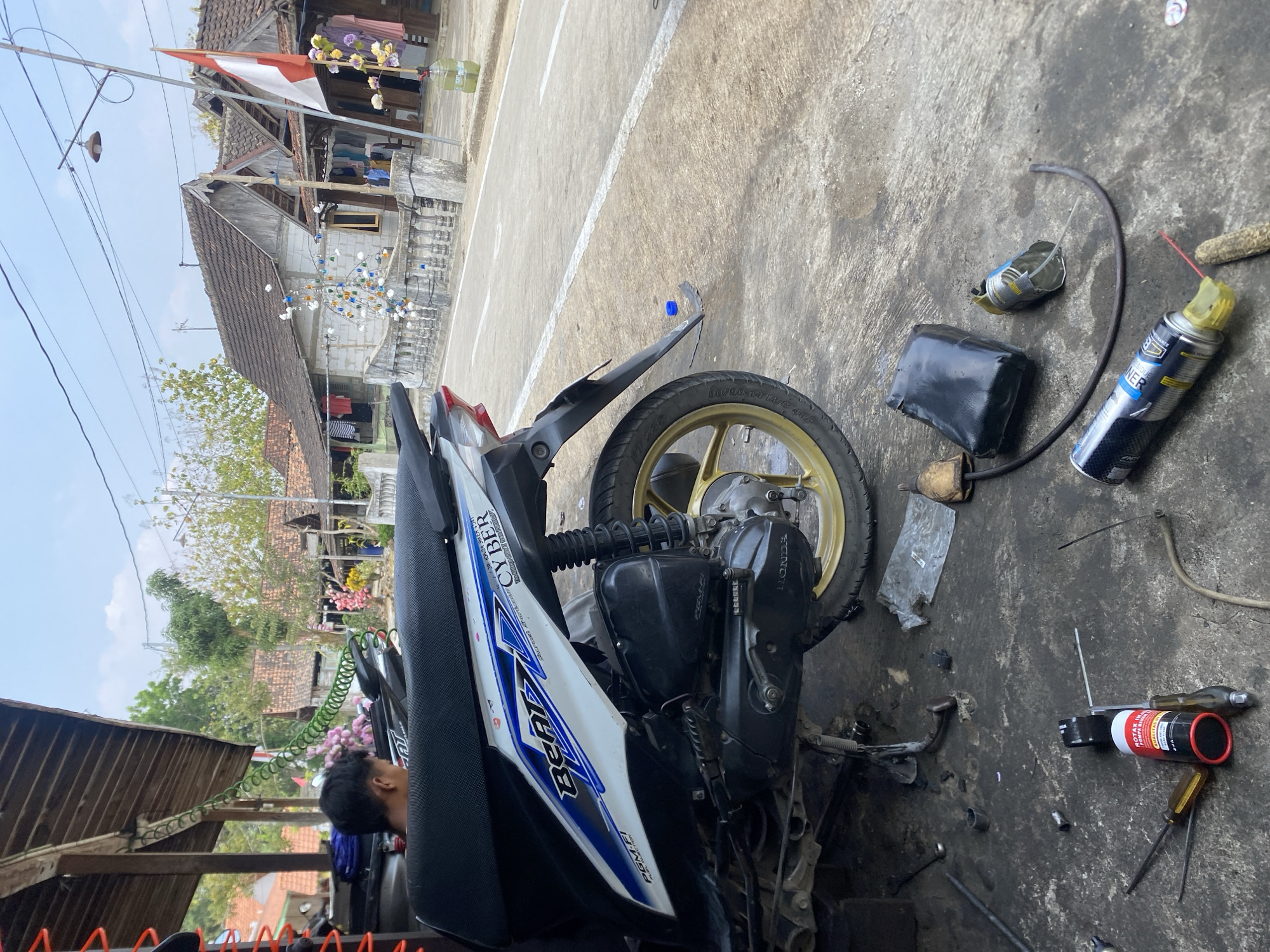 The image depicts a street-level view of a motorcycle repair area on a sunny day. A white and blue Honda Beat scooter is parked, its rear wheel and engine visible. The ground is concrete and appears to be stained with oil and dirt, indicating a workspace for vehicle maintenance. Various tools, including screwdrivers, and cans of lubricant or cleaning spray, are scattered around the scooter. In the background, traditional Indonesian houses with tiled roofs line a paved road. A flag with red and white stripes, possibly the Indonesian flag, is visible on a pole near one of the houses. The overall scene suggests a rural or semi-urban setting with a focus on motorcycle repair.