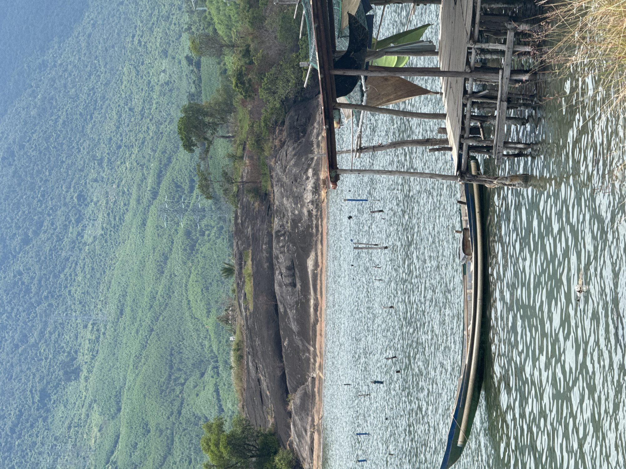 This image depicts a serene waterfront scene, likely located in or near Huế, Vietnam. In the foreground, a narrow, dark-colored boat with a green stripe along its waterline floats on gently rippling water. To the right, a rustic wooden pier juts out into the water, supported by numerous weathered wooden posts. The pier is partially covered by a corrugated metal roof and draped with various pieces of fabric, including a green tarp and a tan cloth. The water also contains several scattered wooden poles, possibly remnants of a fishing structure or markers.
In the mid-ground, a large, dark rock formation dominates the landscape, rising from the water's edge. The rock has an uneven, textured surface, with hints of reddish-brown soil visible in some crevices. Beyond the rock formation, the shore is lined with sparse vegetation and trees.
The background is characterized by a majestic, verdant mountain covered in dense foliage, stretching as far as the eye can see. The mountain slopes are textured with the patterns of numerous trees. Two tall, latticed electricity pylons are visible on the mountainside, indicating the presence of human infrastructure within the natural landscape. The sky above is a pale, hazy blue. The overall atmosphere is tranquil and natural, with elements of both rural life and the imposing beauty of the landscape.