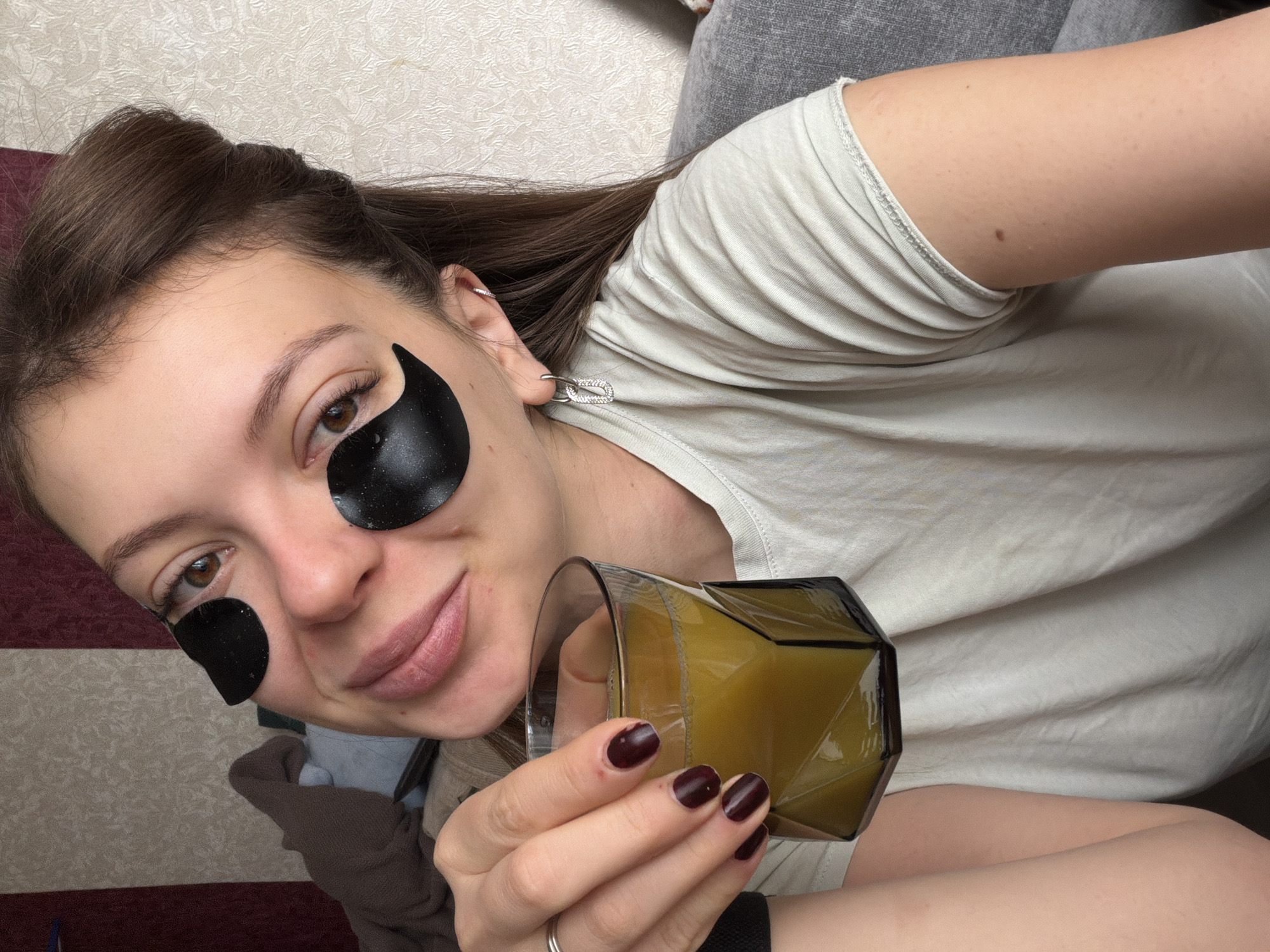 A young adult woman is pictured in a close-up, eye-level shot, reclining indoors. She has long brown hair and brown eyes, with a fair complexion, and is wearing a light-colored, short-sleeved t-shirt. Her facial expression is relaxed, with a gentle smile. She is engaged in a self-care routine, wearing black under-eye patches beneath both eyes.
In her left hand, which has dark red/burgundy nail polish, she holds a clear glass with a distinctive geometric, faceted design, containing a yellowish-green beverage. A simple silver ring is visible on her left ring finger, and a black wristband is on her left wrist. She wears two silver earrings in her right ear: a dangling, rectangular piece adorned with small stones in her lobe, and a smaller hoop in her upper cartilage.
The background consists of a textured wall with vertical stripes in alternating shades of maroon and light beige/off-white. To her right, a grey fabric surface, likely part of a sofa or bedding, provides a soft backdrop. The overall scene conveys a sense of relaxation and comfort within a domestic setting in Khimki, Russia. There is no visible text in the image.