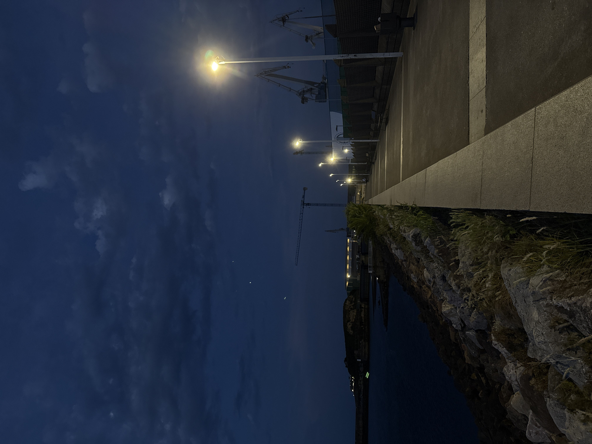 The image depicts a nighttime scene along the coast in Gijón, Spain. A paved promenade runs alongside a rocky embankment bordering the dark water on the left. Streetlights illuminate the promenade, casting a warm glow on the asphalt path. In the distance, across the water, is a coastline with some structures and lights. The sky is a deep blue with scattered clouds, and several points of light, possibly stars or distant aircraft, are visible in the upper sky. Industrial cranes are prominent in the background, suggesting a port or industrial area. The scene is devoid of people, and the overall atmosphere is quiet and serene.