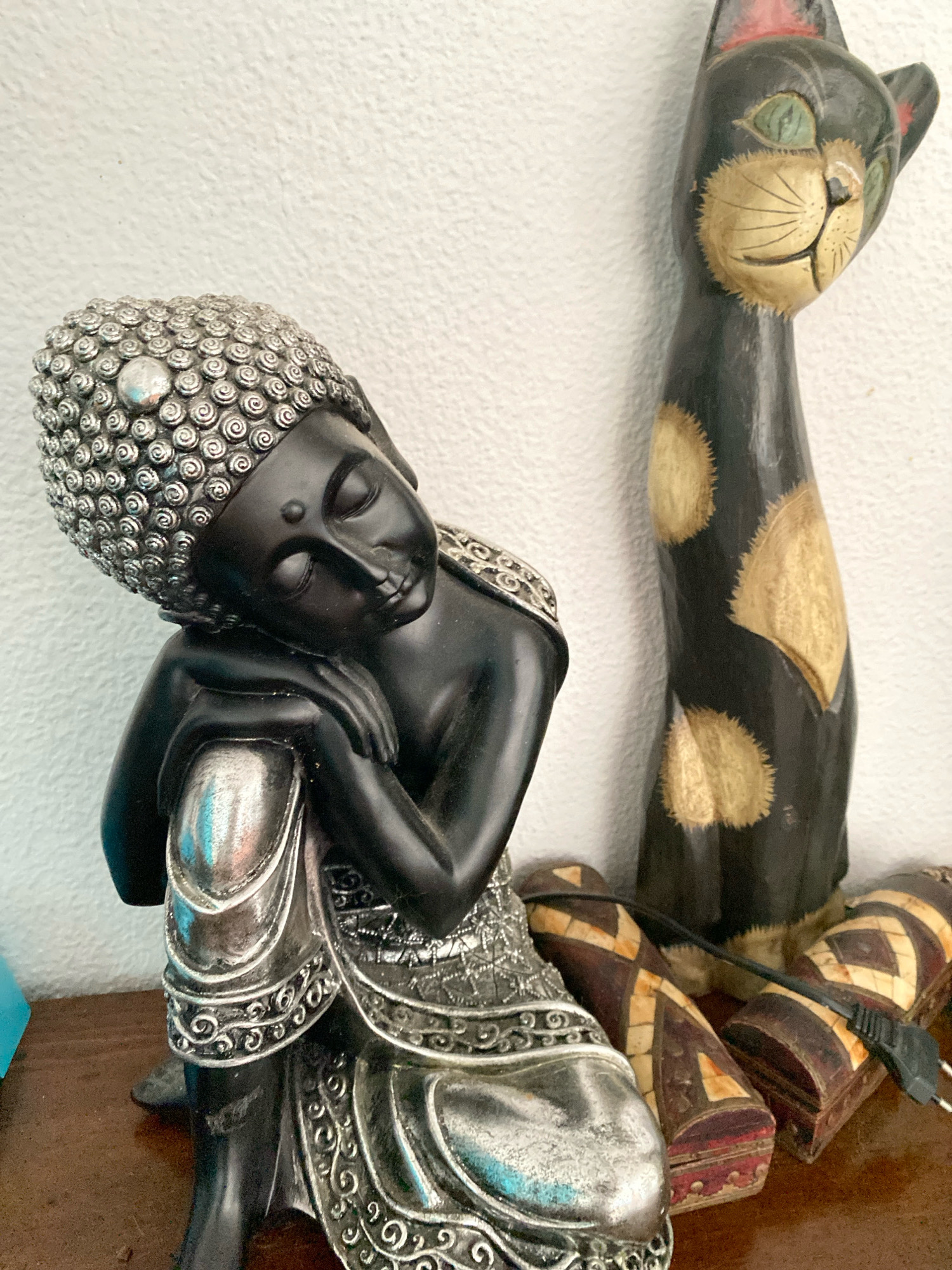 The image displays a close-up of decorative objects on a wooden surface against a textured white wall. The primary subject is a black statue of Buddha in a resting pose, with intricate silver detailing on its hair, clothing, and jewelry. To the right of the Buddha statue is a tall, stylized wooden carving of a black cat with green eyes and light brown spots. In the foreground, partially obscured by the Buddha statue and the cat, are two small, cylindrical wooden boxes with a pattern of alternating dark and light sections. A black electrical cord is visible plugged into one of the boxes. The scene appears to be an indoor setting. The location is identified as Palmela, Portugal.