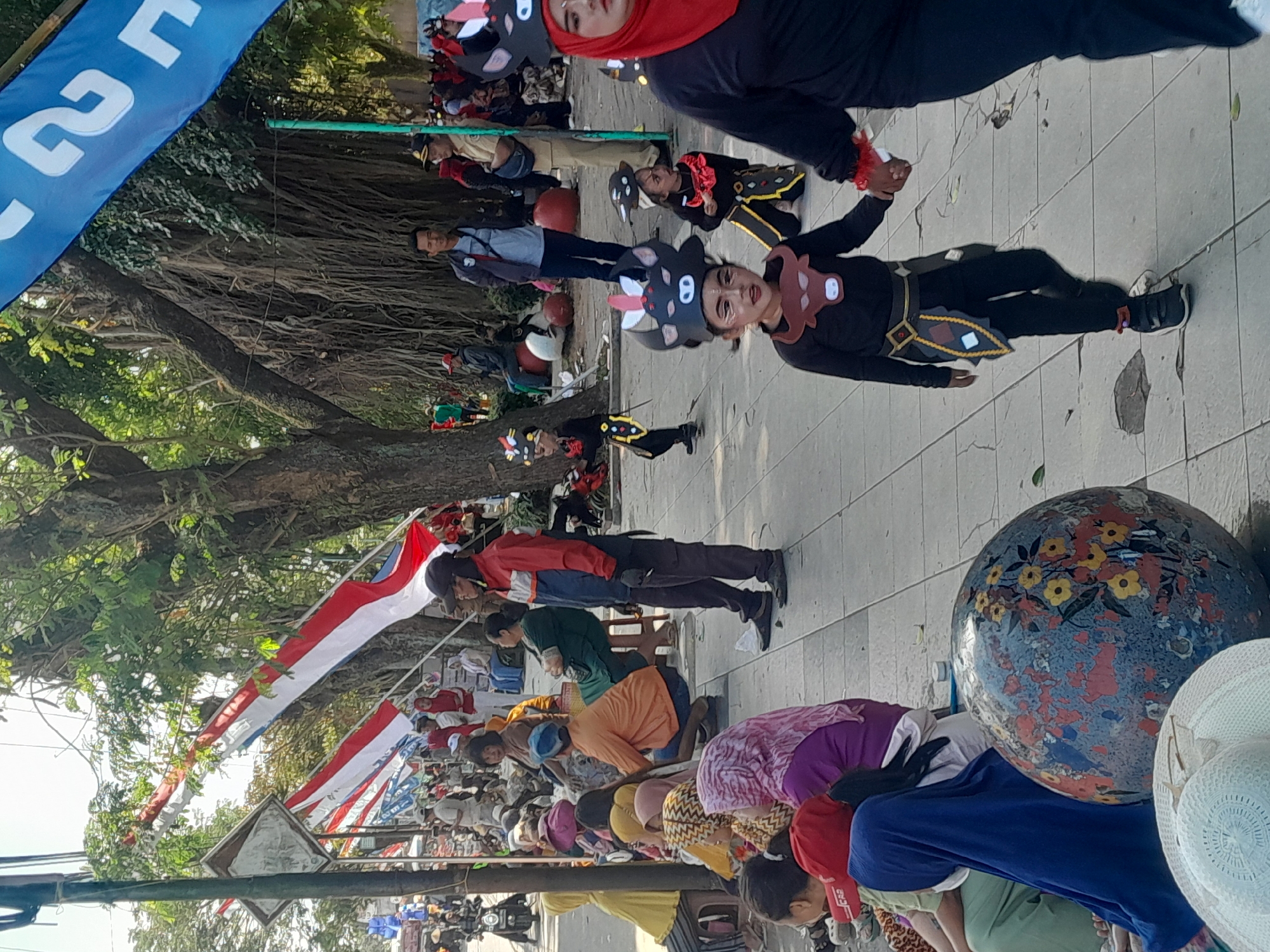 The image depicts a street scene in Genteng Kulon, Indonesia, likely during a festive occasion or parade, with a prominent display of Indonesian flags. In the foreground, a group of children dressed in costume, possibly for a performance or parade, are visible. One child, in focus, is wearing a black outfit with a bull mask and face paint. They are holding hands with an adult also in costume, wearing a red hijab and a black outfit. Another child in a similar costume is seen further back on the right.
To the left of the children, people are seated or standing, some observing the events. One person is wearing a red and blue jacket, standing with hands clasped, and holding a cigarette. Further to the left, a large decorative globe with floral patterns is visible, along with straw hats.
The background features a street lined with trees, some with exposed aerial roots, indicating the presence of large, mature trees. Several Indonesian flags, red and white, are strung up along the street, fluttering in the breeze. A large blue banner with white text is also visible in the upper right corner. Other individuals in civilian attire, including men in uniforms, are scattered throughout the scene, suggesting a public gathering. The ground is paved with light-colored tiles. The overall atmosphere appears to be that of a community event or celebration.