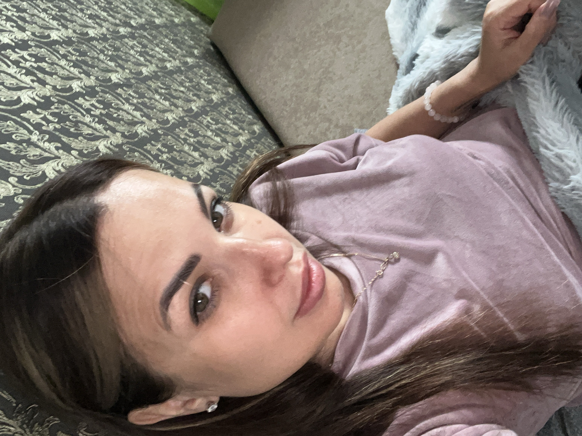 The image shows a close-up, high-angle shot of a young woman lying on a sofa. She is wearing a light purple, long-sleeved shirt. A light gray, fuzzy blanket is draped over her right arm. She has long, dark brown hair that is spread out around her. Her left arm is visible, and she is wearing a bracelet made of small, light pink beads. She is looking up and slightly to her left, with a neutral expression. Her makeup includes dark eyebrows and eyeliner, and her lips are a rosy pink color. A delicate necklace with a small pendant is visible on her shirt. The sofa appears to be covered in a patterned dark fabric, and a plain beige cushion is visible behind her head. The lighting suggests it could be daytime, but the exact time is not clear. No text is visible in the image.