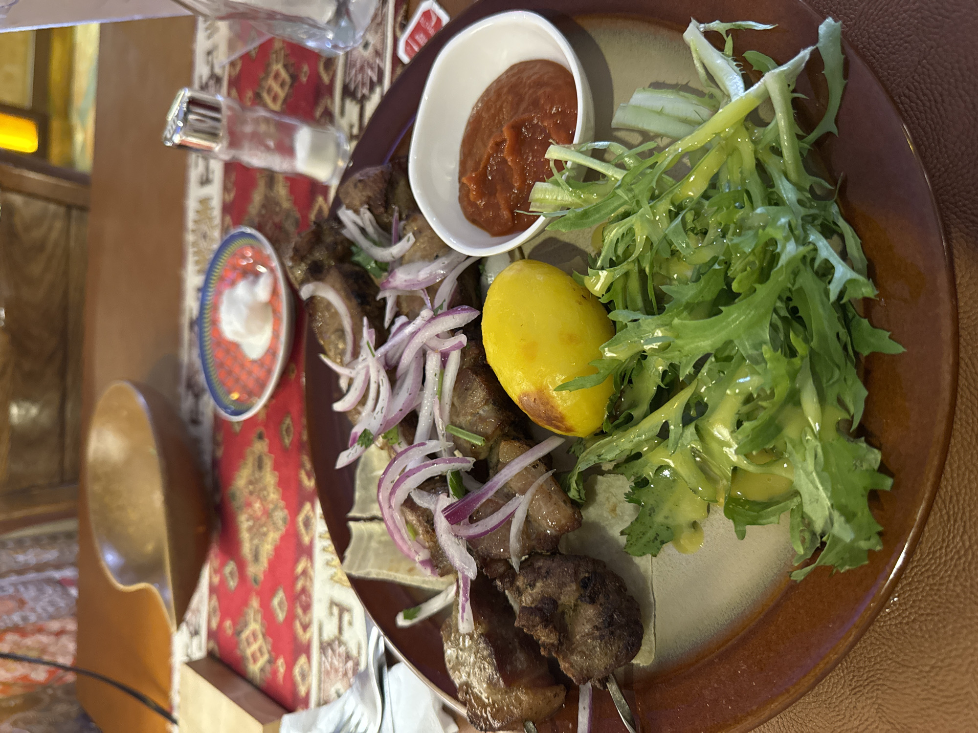 The image depicts a plate of food, likely taken in a restaurant setting, given the presence of tableware and a decorative carpet on the table. The main dish appears to be shashlik (grilled meat skewers) garnished with red onion slices and served with a side of boiled potato, a small bowl of red sauce, and a salad with green leaves, possibly a mix of lettuce and herbs, drizzled with a light-colored dressing. A salt shaker is visible in the background, along with a small dish containing a white sauce. The presence of a decorative rug on the table suggests a culturally themed or traditional establishment. The overall impression is that of a meal served in a restaurant, with a focus on grilled meat and accompanying side dishes. It is not possible to confirm its location in Ulaanbaatar, Mongolia, without additional contextual information.