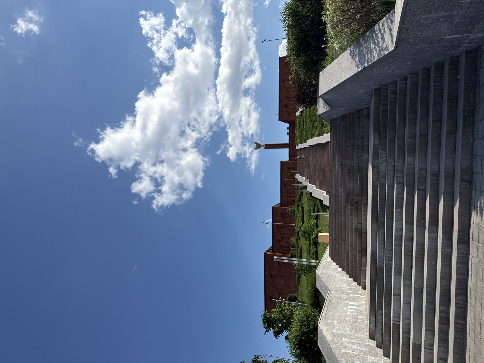 The image shows a view of the Kazan Kremlin from a lower vantage point, likely a nearby park or plaza, in Kazan, Russia. The Kremlin's red brick walls are visible, topped with green foliage and interspersed with modern lampposts. A prominent gold spire with a crescent moon adorns one of the Kremlin's towers. A set of concrete stairs lead up and out of frame on the right. The sky is a clear blue with scattered cumulus clouds on the left side of the frame.
