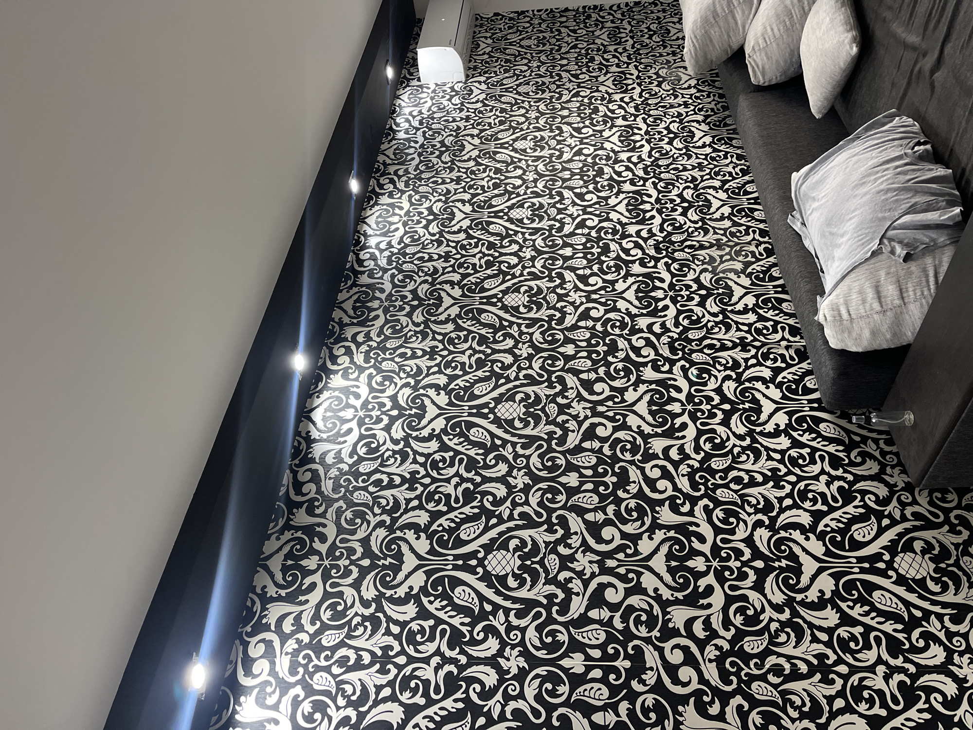 This is a medium-shot, eye-level interior photograph of a living room in Stavropol, Russia. The dominant feature is a black and white patterned wallpaper that covers the entire wall. The pattern is intricate and consists of swirling, floral, and possibly avian motifs. A black soffit with integrated LED lighting runs along the top of the wall, casting a bright, cool light downwards onto the wallpaper. To the right, a white air conditioning unit is mounted high on the wall, partially visible. In the foreground, a dark grey sofa is visible, with several light grey pillows and one pillow covered with a light grey t-shirt. The t-shirt is draped loosely over the pillow. A small, clear glass bottle is placed on a dark surface to the right of the sofa. The room appears modern and somewhat minimalist, with the wallpaper providing a strong decorative element.