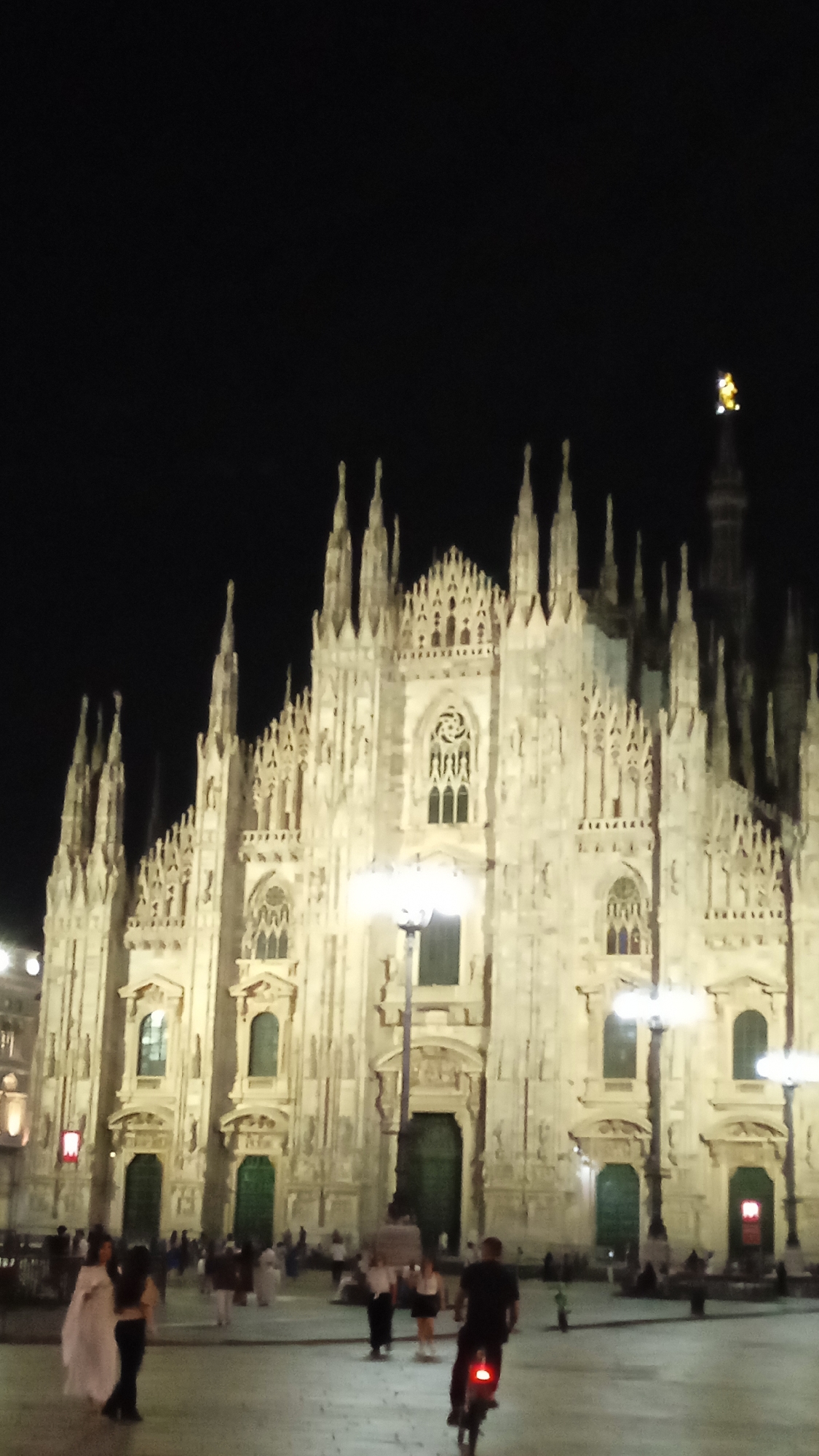 The image captures the facade of the Milan Cathedral (Duomo di Milano) at night. The cathedral's intricate Gothic architecture is illuminated, highlighting its numerous spires and detailed carvings. In the foreground, the square in front of the cathedral is populated by several individuals. Two people, one wearing a light-colored garment and the other in dark clothing, are standing together on the left. Further back, a group of people are scattered throughout the plaza. In the center of the image, a person is riding a bicycle, their red taillight visible against the dim lighting. Streetlights on tall poles are positioned in front of the cathedral, contributing to the overall illumination of the scene. The sky above is dark, indicating it is nighttime.