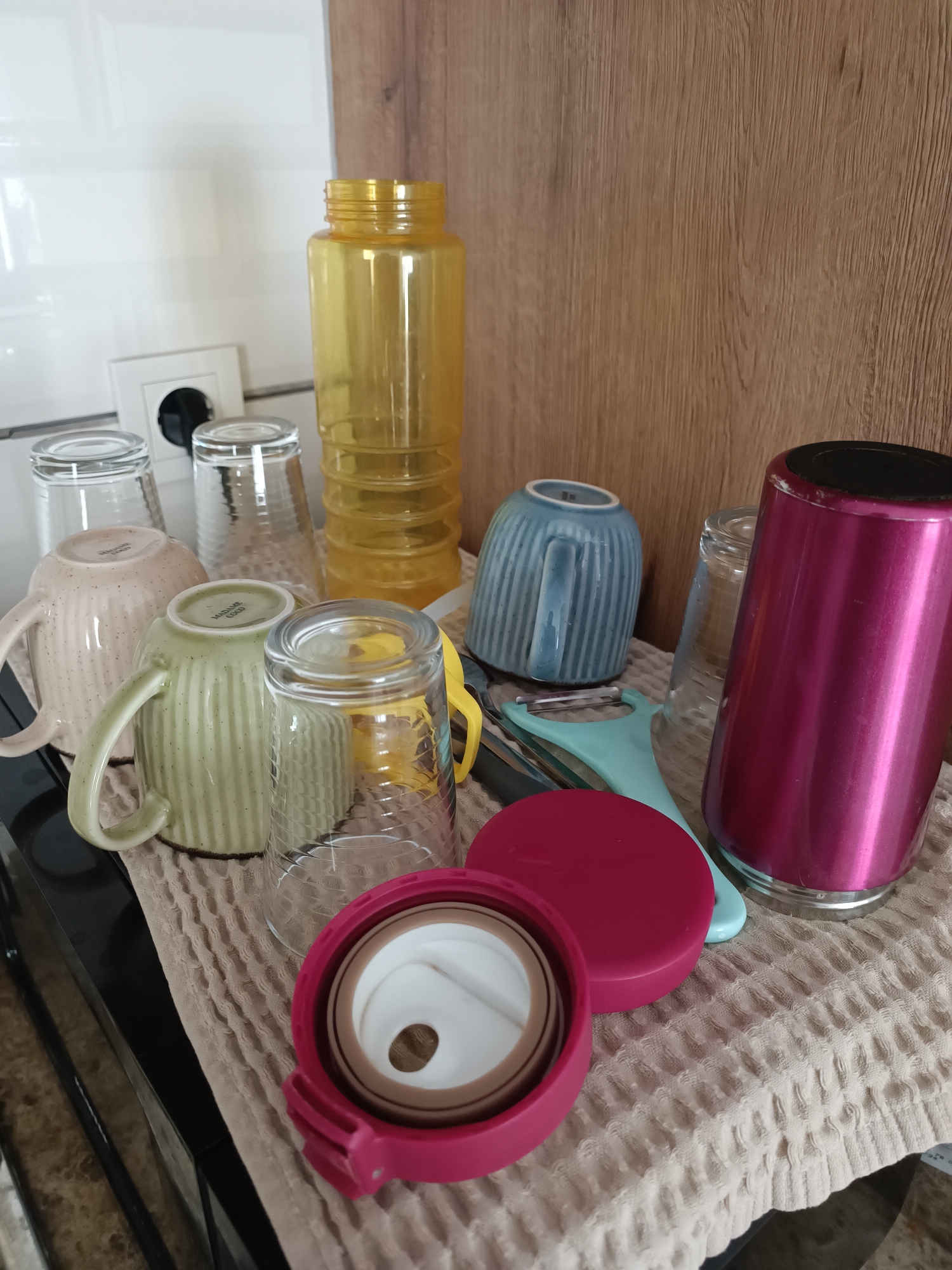 A collection of kitchenware, including cups, glasses, a yellow plastic bottle, a pink thermal mug, and a peeler, is arranged on a textured beige cloth. The items are positioned on a dark countertop, likely in a kitchen setting. In the background, a white tiled wall with a power outlet and a wooden cabinet are visible. The overall scene appears to be an indoor still life. The image does not provide any information that could place it in Silifke, Turkey, or any other specific city.