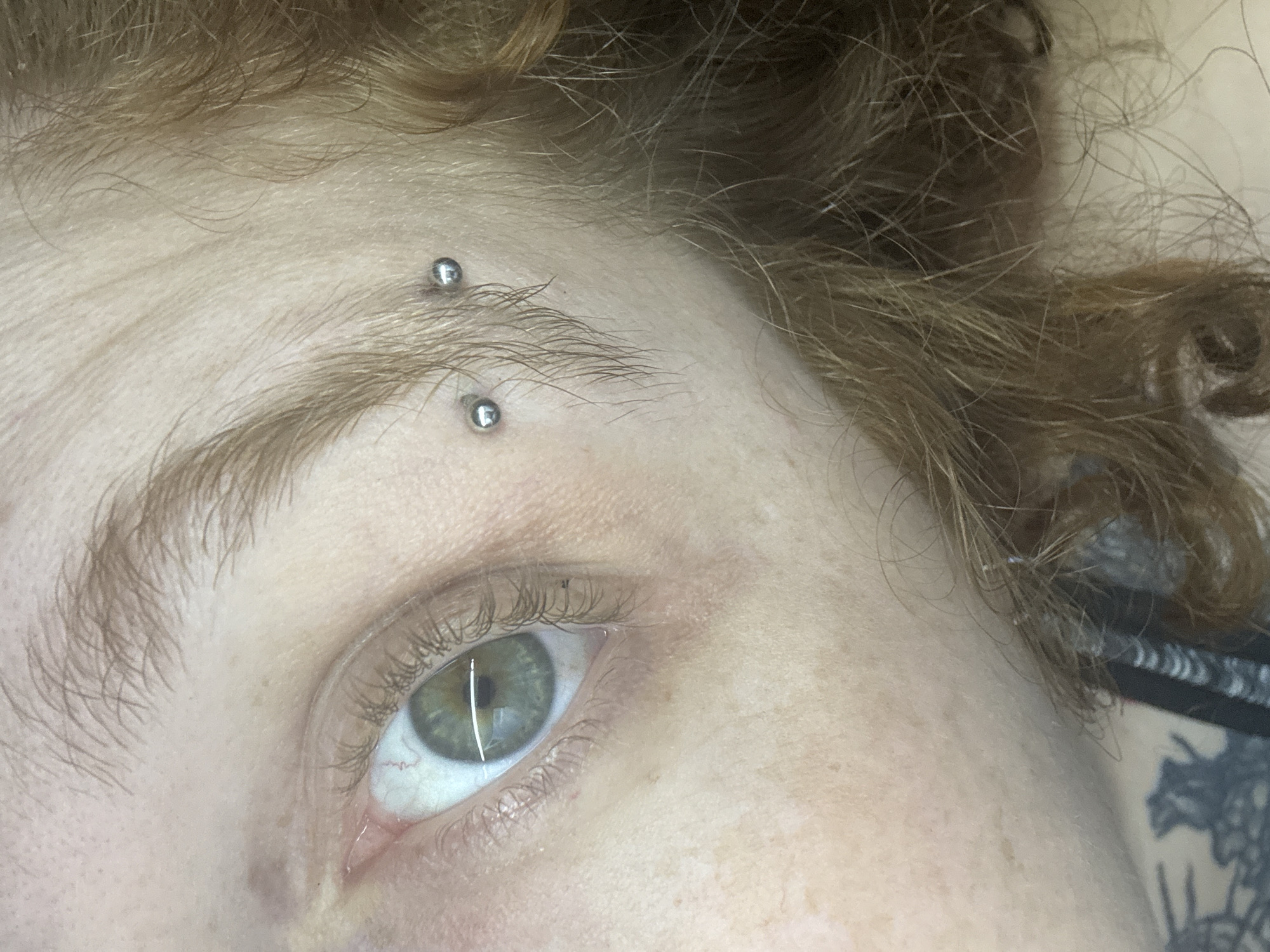 This close-up image, taken in Buenos Aires, Argentina, captures the right eye and eyebrow of a person. The subject has fair skin with subtle freckles. Two vertical barbell piercings, each with small silver spheres, are visible through the eyebrow. The eye features a green or hazel iris, with visible eyelashes and some faint red veins in the sclera. A prominent vertical reflection of a light source is seen in the pupil. Reddish-brown, curly hair frames the forehead and is also visible on the right side of the frame. On the far right, a dark, intricate tattoo, possibly depicting floral or abstract patterns, is partially visible beneath the hair. The image is a static portrait, focusing on these facial details. No specific activity or visible text is present.