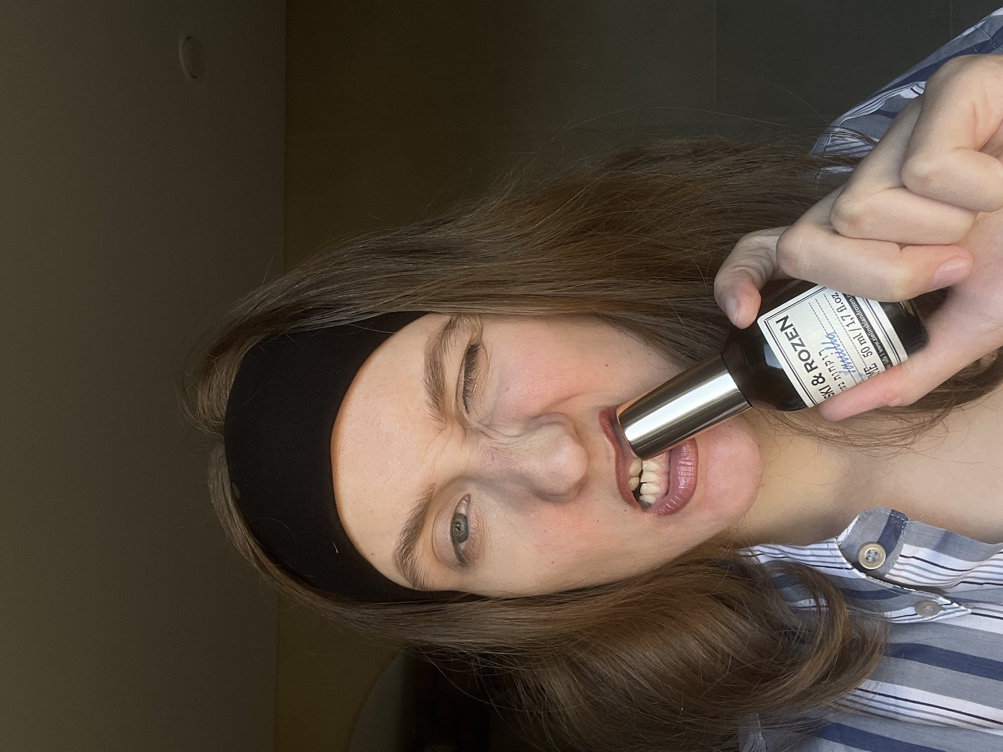 A woman with light skin, brown hair, and a black headband is holding a bottle of "SKI & ROZEN Vanilla ME" in her teeth. She is wearing a blue and white striped shirt. The background appears to be an interior wall. It is not possible to determine the location in the city of Подшивалово, Russia, or any connection to it, from the image.