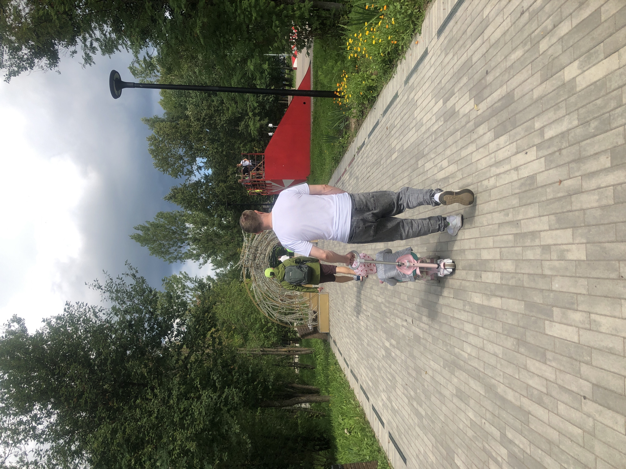 The image shows a man walking away from the camera on a paved pathway in a park. He is wearing a white t-shirt and grey jeans. In front of him, a young child is sitting on a pink tricycle with a parent pushing it. The pathway is lined with trees and grass. In the background, there is a red ramp structure, possibly for skateboarding or BMX biking, with several people on top. A lamppost is visible on the right side of the image. The sky is cloudy. The scene appears to be in a public park within the city of Tutaev, Russia, likely a recreational area.