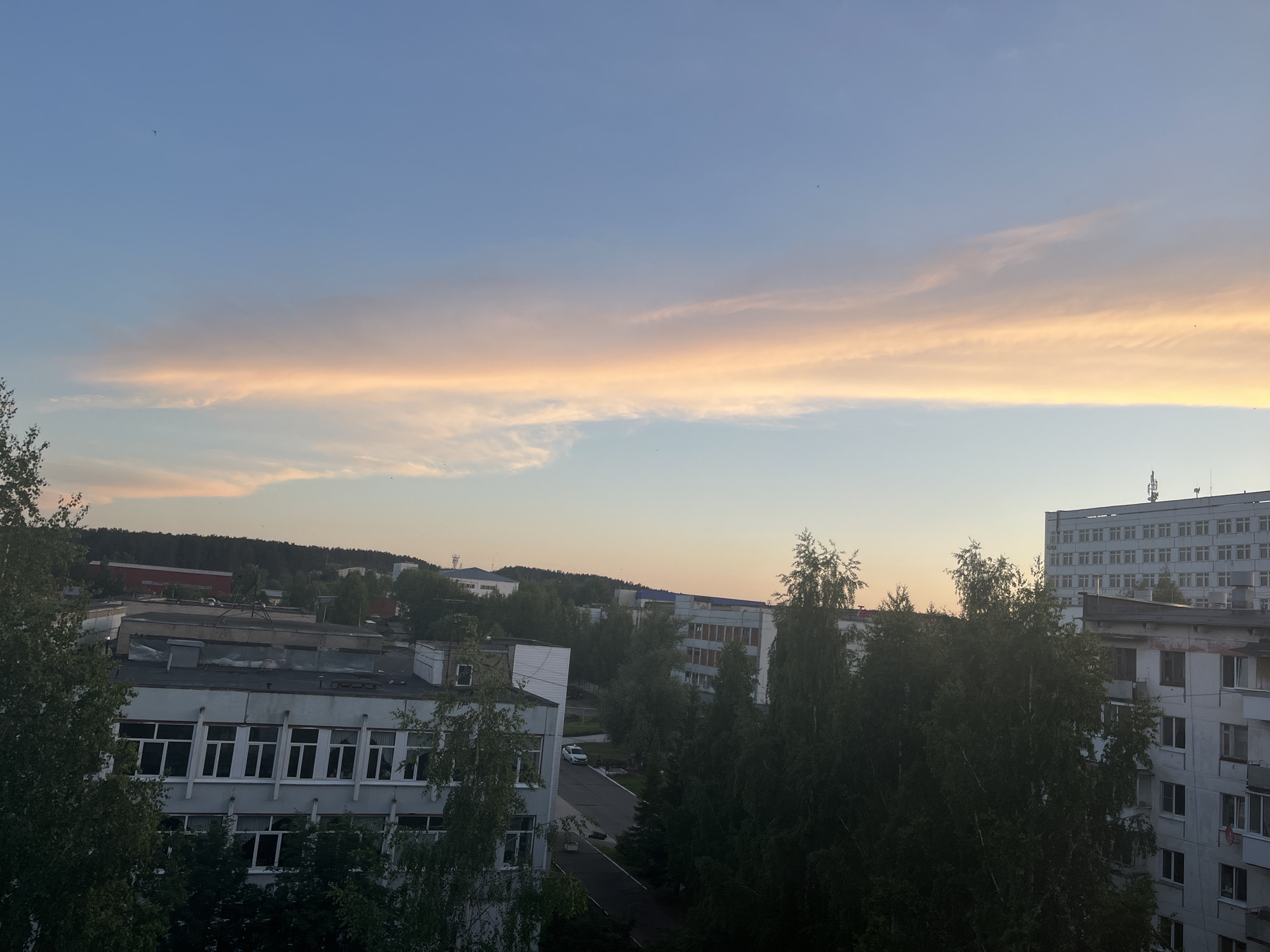 This elevated shot captures a panoramic view of a town, likely Chaikovsky, Russia, during either a serene sunrise or sunset. The sky dominates the upper half of the image, showcasing a beautiful gradient of colors. A deep blue occupies the top, gradually transitioning to a lighter blue and then a prominent, elongated band of soft orange, pink, and yellowish clouds stretching horizontally across the mid-sky. Below this colorful cloud formation, the sky lightens to a pale blue with some faint, wispy white clouds. Several tiny dark specks, probably birds, are visible against the blue expanse.
The lower half of the frame reveals an urban landscape nestled amongst abundant greenery. In the immediate foreground and mid-ground, numerous dark green trees, possibly birches given their lighter bark in places, create a dense canopy that partially obscures buildings. Architectural subjects include a long, multi-story building with numerous rectangular windows on the left, possibly an administrative or educational facility. Further back and to the right, a tall, light-colored apartment block with many windows stands prominently. Other buildings are scattered throughout, blending into the landscape. A paved road or pathway winds between some structures, with a white car visible on it. In the distant background, dark forested hills or a dense treeline stretch across the horizon, suggesting the town is surrounded by nature. No specific human activity is distinctly visible, and no text is apparent on any structures. The overall mood is calm and picturesque, highlighting the interplay of urban development and natural beauty under a captivating sky.