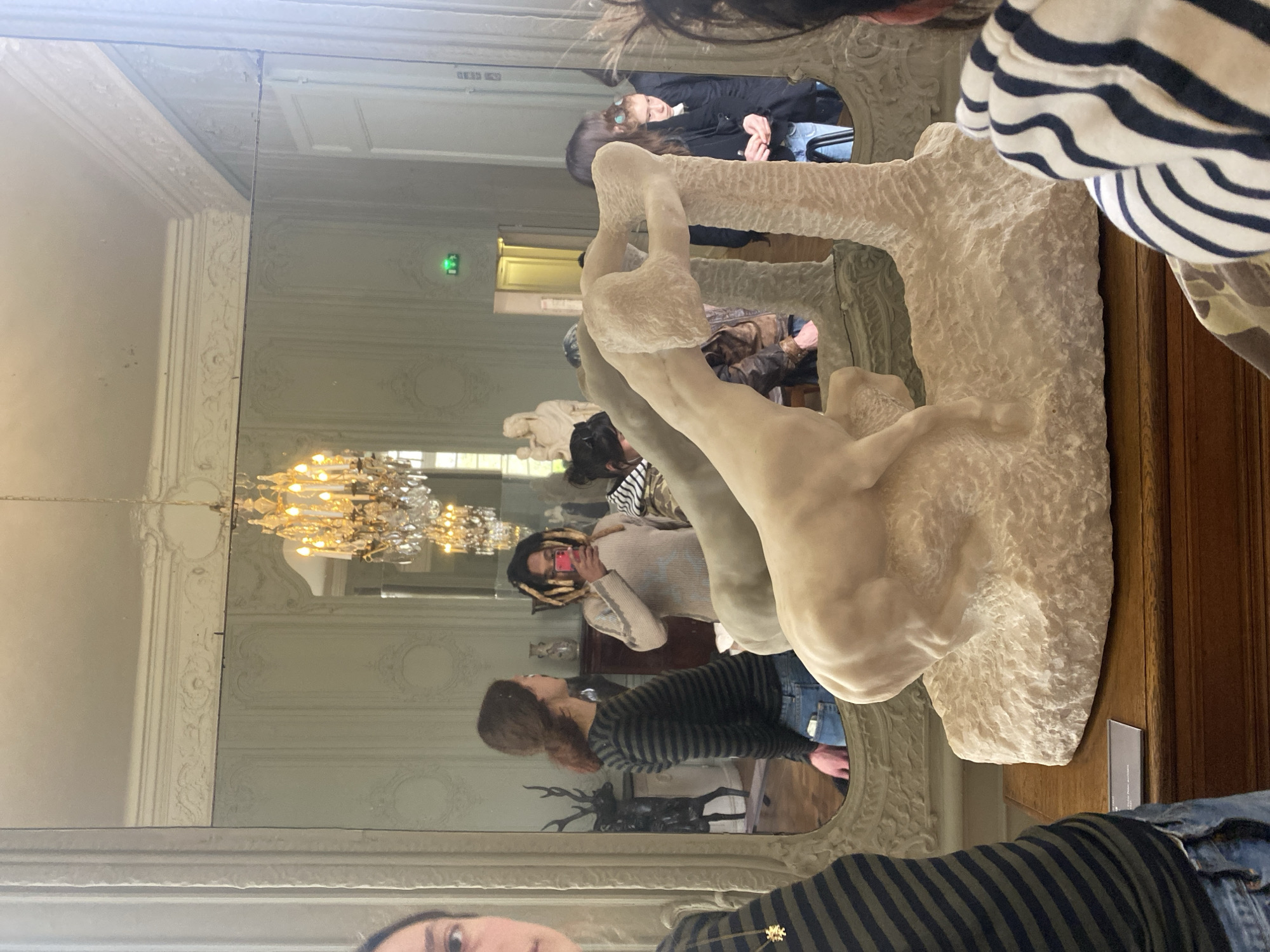 The image depicts an interior scene within the Musée Rodin, located in Paris, France. The primary subject is a cream-colored, stylized sculpture of a dynamic, possibly rearing, horse-like figure, resting on a textured base atop a dark wooden display cabinet in the foreground. A small, angled informational plaque is visible on the cabinet in front of the sculpture.
The room features light-colored, ornate walls with classical French paneling and intricate moldings. A large, decorative mirror with a carved frame dominates the background, reflecting various elements of the room and its occupants.
Reflected in the mirror are several individuals and artworks. Towards the left of the reflection, a female with long brown hair in a ponytail, wearing a dark green and black striped long-sleeved shirt and blue jeans, is seen from the back. In the center-right reflection, a person with light-colored dreadlocks or braids, wearing a light beige long-sleeved top, holds a pink-cased smartphone up, appearing to photograph the scene. Further to the right in the reflection, a young female with dark hair, clad in a dark coat and blue jeans, is partially visible, looking downwards.
Additional reflected elements include a grand chandelier with multiple lit bulbs, a dark sculpture resembling a stag or deer with prominent antlers, and a classical white marble statue, possibly depicting a mother and child. A doorway with a yellow or golden frame and a green exit sign is also visible in the reflection.
In the immediate foreground, to the left, the partial view of a person's left side is visible, wearing a dark green and black striped shirt (matching the reflected individual) and blue jeans, with a gold star-shaped pendant on a chain. To the immediate right in the foreground, the back of a person's head and shoulder are visible, wearing a garment with black and white horizontal stripes.
