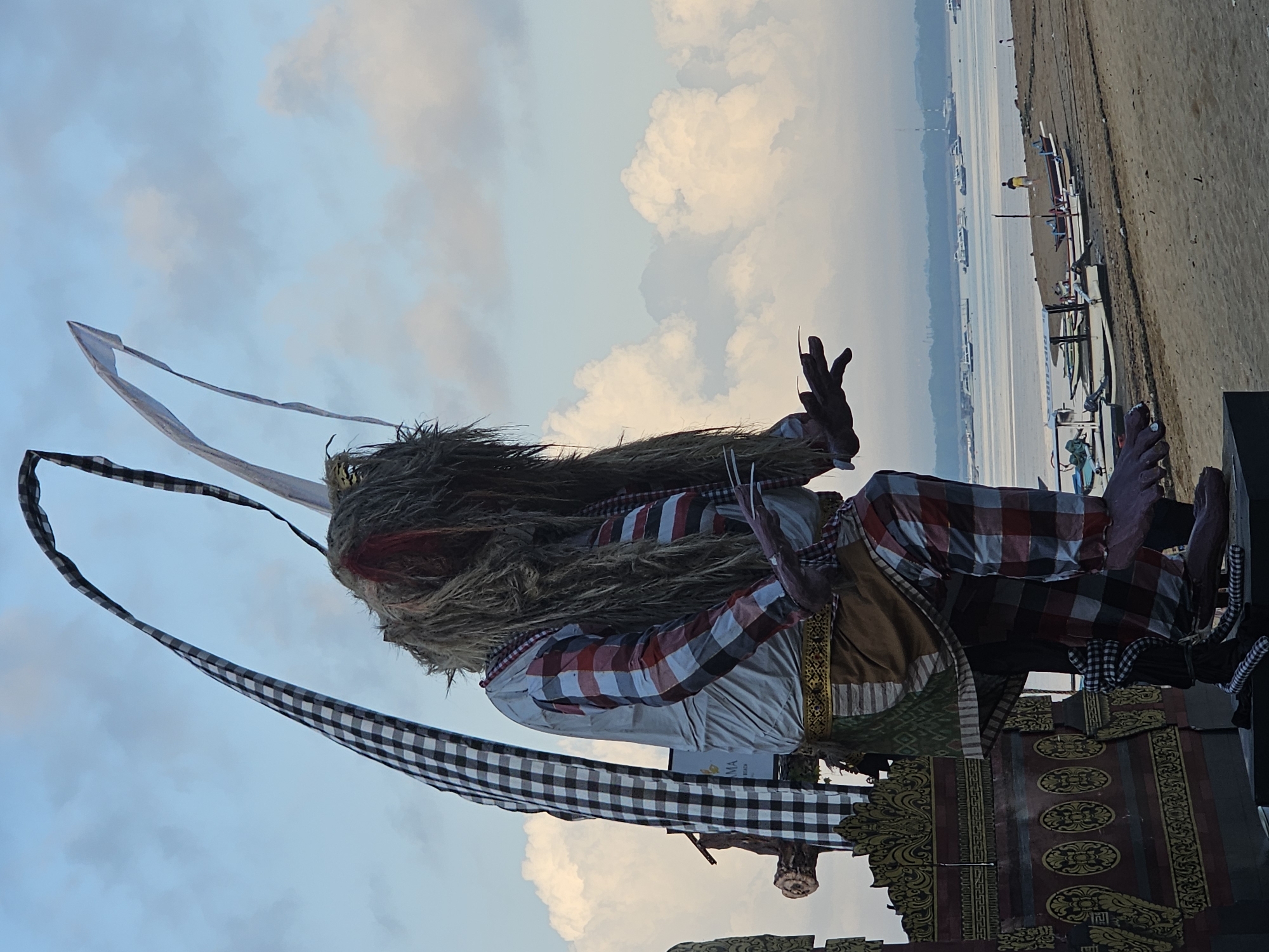 A large, effigy of a mythical creature, possibly a demon or ogre, stands on a beach with its torso angled to the right. The effigy is dressed in a red and white checkered long-sleeved shirt and pants. It has long, straw-like hair and red markings around its eyes. Its hands are outstretched, with long, sharp fingernails. The effigy is adorned with a patterned sash and a black and white checkered cloth draped over its left shoulder. Behind the effigy, there is a decorative structure with ornate carvings. The background shows a sandy beach with several small boats pulled ashore, and a calm body of water with numerous boats in the distance. The sky is cloudy with patches of blue. The scene appears to be in Denpasar, Indonesia, possibly during a cultural event or festival given the nature of the effigy. There is one person visible in the distance walking on the beach.