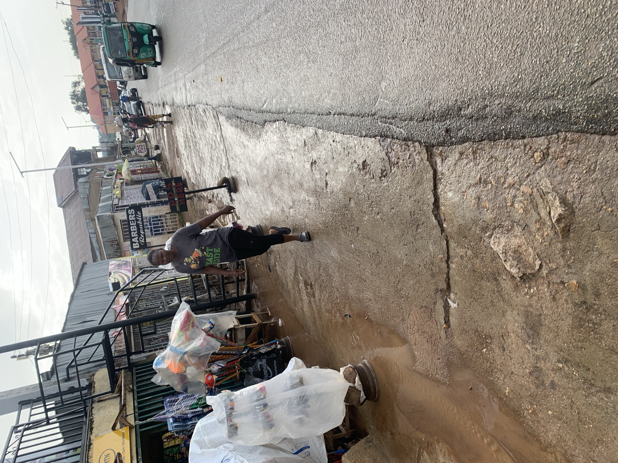 This street-level photograph captures a wet and active urban scene in Gura Top, Nigeria, likely during daytime under overcast skies. The asphalt road, stretching into the distance, is visibly damp and features puddles, indicating recent rainfall.
In the foreground, on the right side of the image, a person wearing a grey t-shirt with "EAST MODE" printed on it and dark trousers is walking towards the viewer, looking directly at the camera. Their left arm is slightly extended. The unpaved ground adjacent to the road is rough, muddy, and uneven, with standing water. Scattered along this makeshift shoulder are various plastic-wrapped items and two discarded vehicle wheel rims.
On the left side, a series of small shops and market stalls line the street. Notable signs include a black one reading "BARBERS Republic" and a white sign displaying "POS HERE." A yellow sign further down features the "MTN" logo with partial text "Angw" and "Broadband." Many goods, some wrapped in plastic, are displayed outside these establishments. Further down the street, another individual can be seen walking, possibly carrying items on their head.
Vehicles are visible on the wet road in the background, including a green 'keke napep' (motorized tricycle taxi) driving away from the viewer, followed by a white bus or van and other cars. The sky is bright but uniformly grey, suggesting an overcast day. The scene portrays daily commerce and movement within a bustling urban environment under damp conditions.