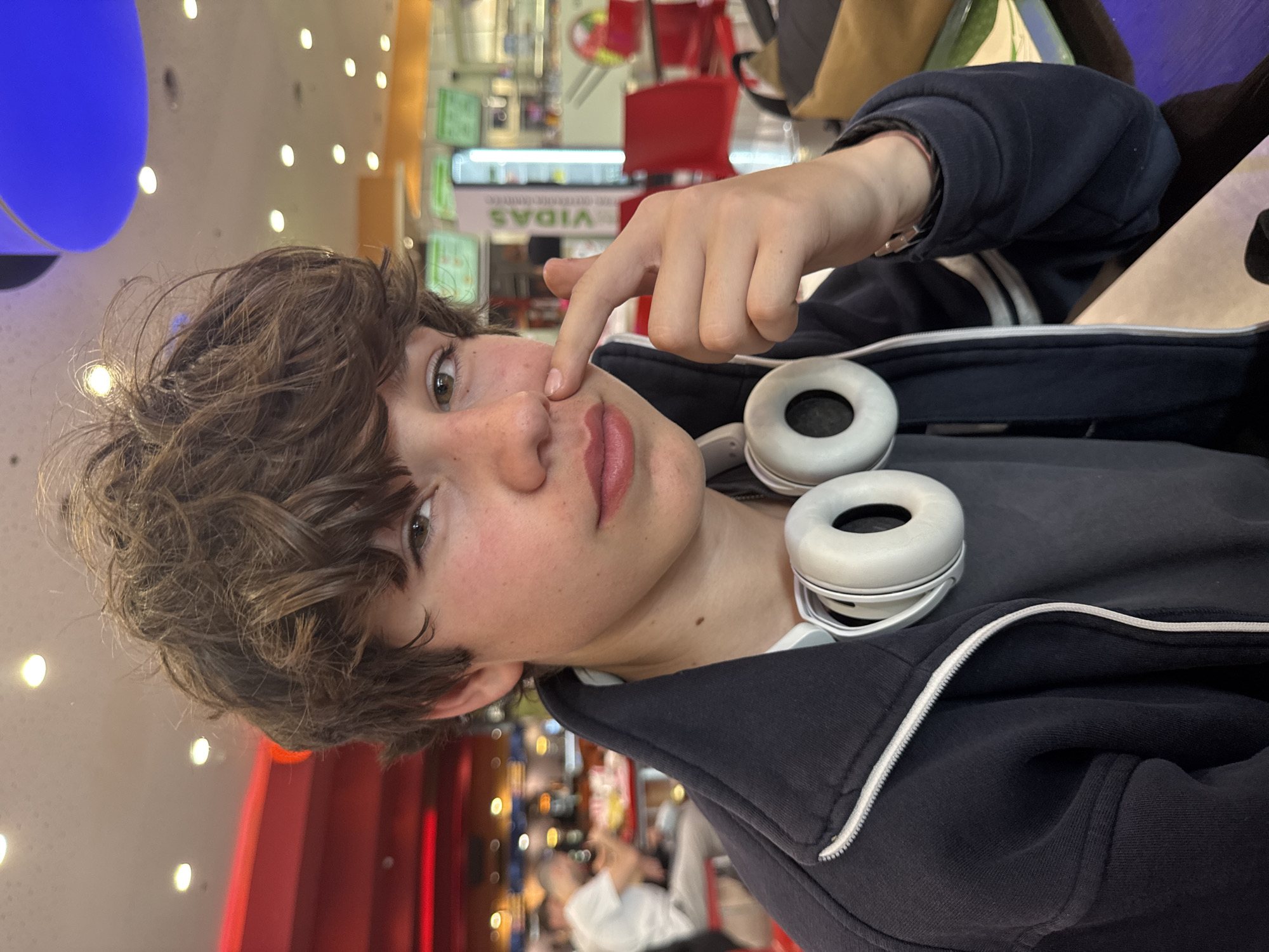 A young person with curly brown hair, wearing a dark zip-up jacket with white stripes, and white headphones around their neck, is shown touching their nose with their finger. The background is a public space with bright lights and store displays, possibly a shopping mall. A blue circular light fixture is visible near the ceiling. The scene appears to be indoors.