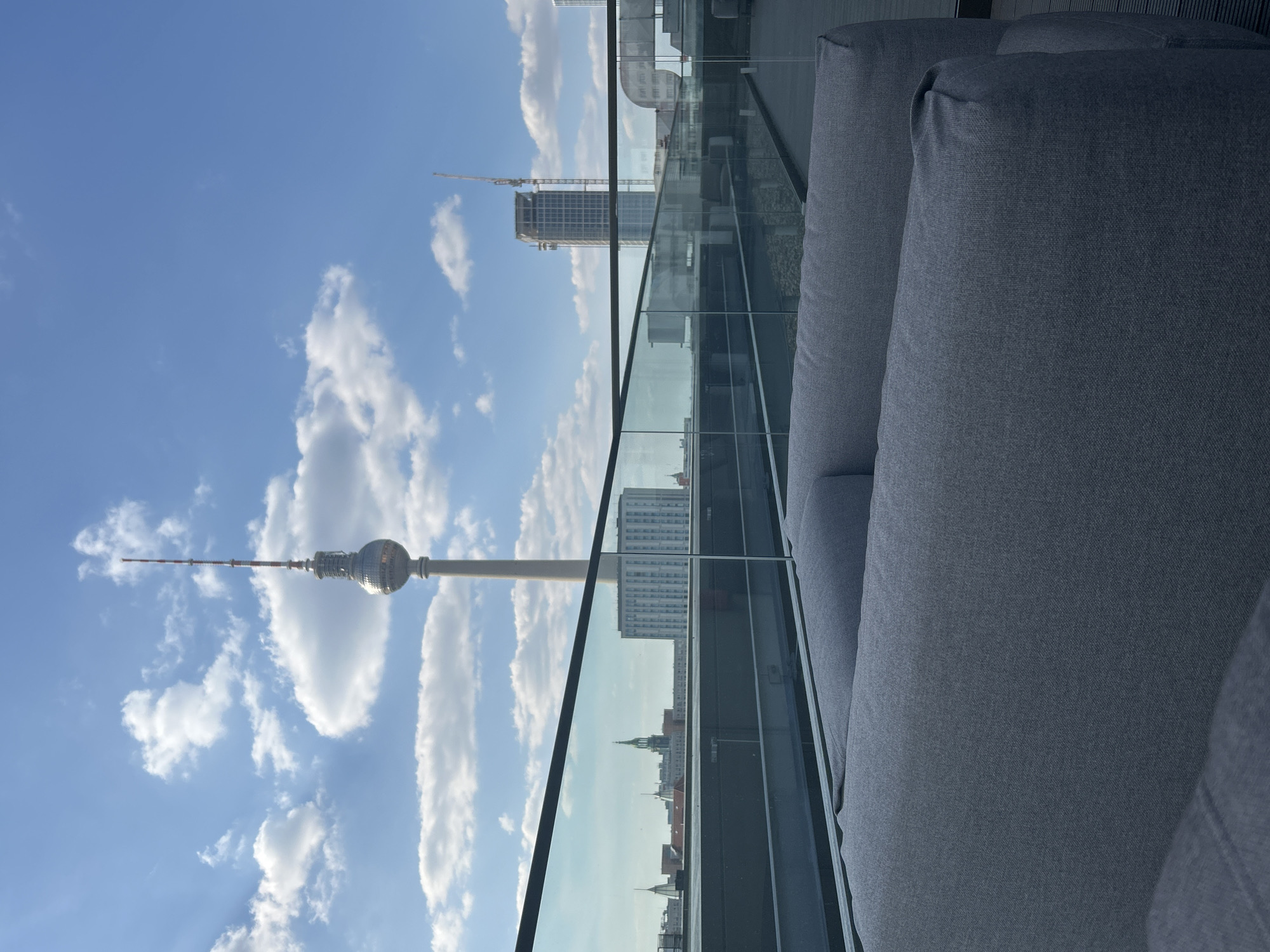 The image shows the Berlin TV Tower as seen from a high-rise balcony in Berlin, Germany. A modern glass railing separates the balcony from the cityscape. A gray outdoor couch is in the foreground. Reflected in the glass railing are city buildings. The sky is blue with scattered clouds. A building under construction with a crane is visible in the distance.