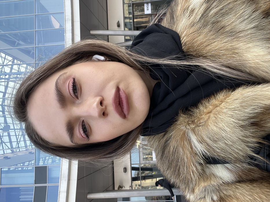 A close-up selfie-style photograph depicts a young adult female looking directly into the camera, positioned outdoors in Saint Petersburg, Russia. She has light brown hair, fair skin, and is wearing pink eyeshadow, mascara, and dark pink lipstick. A white wireless earbud is visible in her right ear. She is dressed in a multi-toned brown fur coat over a black garment, with a black scarf wrapped around her neck.
The background features a modern building characterized by a prominent, curved, and segmented blue-tinted glass facade that reflects the sky and surroundings. Grey architectural pillars are visible at the lower sections of the building. To the left, a distant, dark-clad figure is partially visible, appearing to interact with a building element near an entrance. To the right, a white sign with black Cyrillic text "ВХОД" (ENTRANCE) is displayed on a dark panel next to an entrance, confirming the Russian location. The architectural style, specifically the glass dome, is consistent with buildings such as the Piterland Aquapark and Shopping Mall.