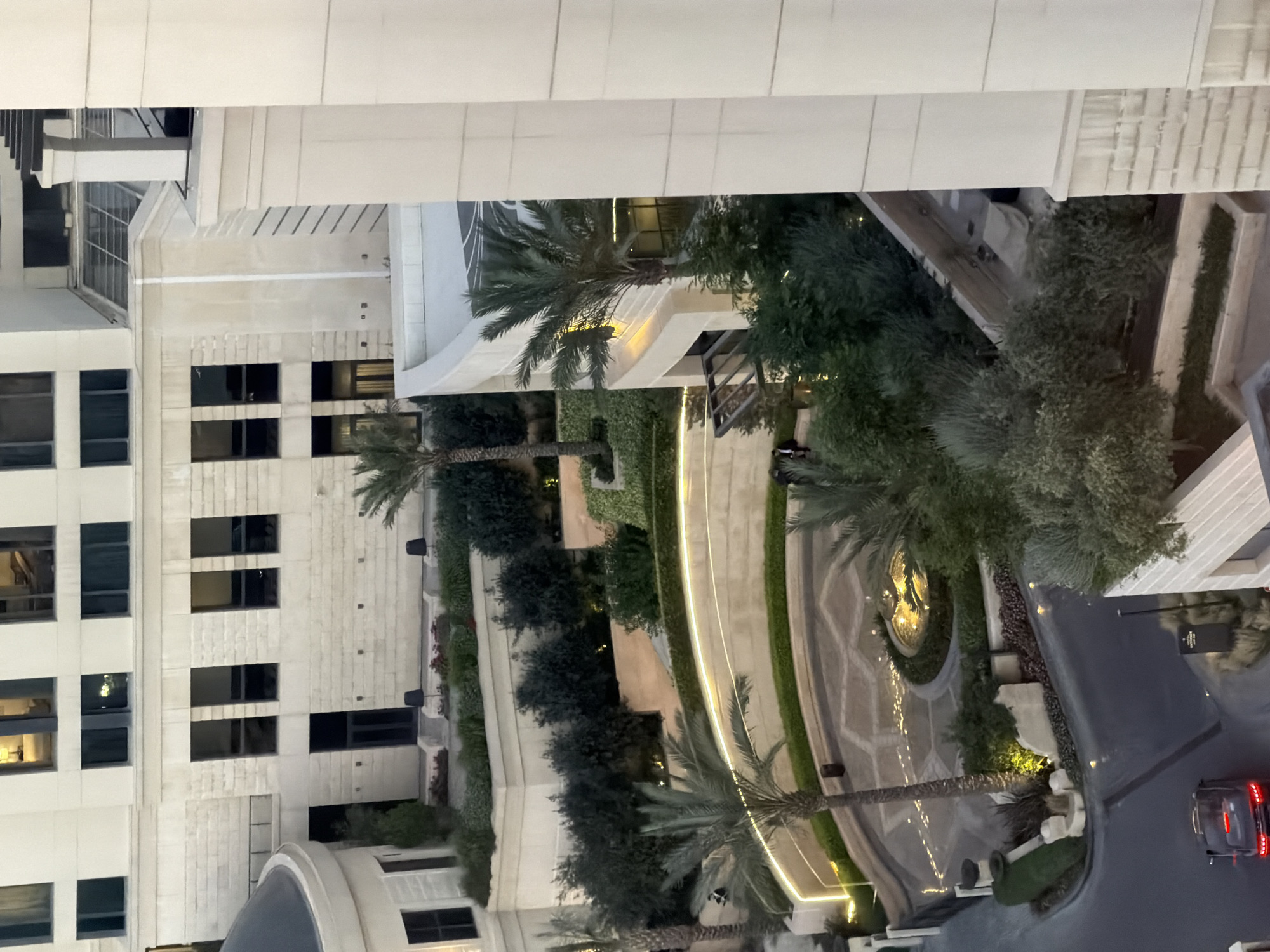 In an aerial view, a luxury hotel complex is depicted, likely the Grand Hyatt Amman, situated in Amman, Jordan. The architectural style features modern stone exteriors with clean lines and numerous windows. The foreground shows a winding driveway leading to a circular entrance area. This entrance is illuminated by warm lighting and features a fountain at its center, surrounded by lush greenery and palm trees.
Two figures, dressed in dark attire, are visible near the entrance, one standing and another seated. A dark-colored luxury sedan is parked on the road in the lower left corner. The surrounding landscape includes manicured gardens, potted plants, and several palm trees, indicating a well-maintained outdoor space. The upper portion of the image reveals more of the hotel's structure, with multiple floors and windows, some of which are illuminated, suggesting indoor activity. The overall impression is one of opulence and sophisticated urban design.