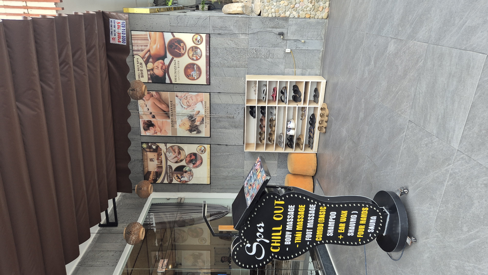 The image shows the exterior of a spa called "Spa Chill Out" in Da Nang, Vietnam. A black, heart-shaped sign with white and yellow lettering lists spa services, including body massage, Thai massage, foot massage, combo treatments, ear wax, shaving, four hands, and sauna. In front of the sign is a shoe rack filled with various pairs of sandals and flip-flops. Three framed posters displaying spa services are mounted on the textured gray stone wall behind the shoe rack. The spa appears to have an awning made of brown fabric. A few orange cylindrical ottomans are placed near the sign. The flooring is composed of large, light gray square tiles.