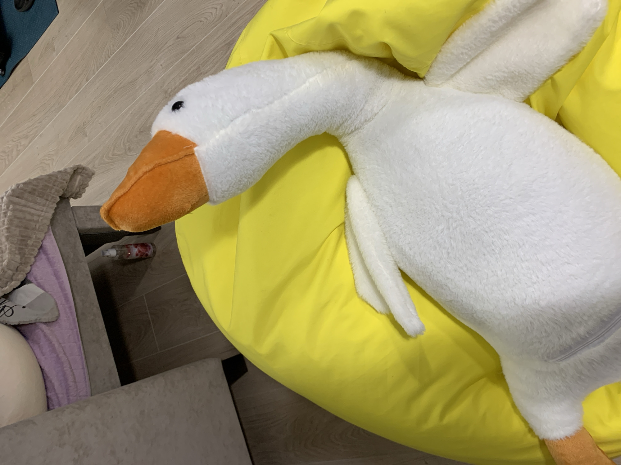 A white stuffed goose with an orange beak and feet is lying on a bright yellow beanbag chair. The goose is positioned diagonally, with its head turned to the left and its body extending towards the bottom right of the frame. The beanbag is large and takes up the majority of the image. In the background, to the left, a portion of a beige sofa and a lavender-colored blanket can be seen. To the right of the sofa, a small, clear bottle with a pink liquid is visible on the wooden floor. The floor appears to be made of light brown wood planks. The lighting suggests an indoor setting, possibly a living room or play area. There are no people visible in the image. The scene is located in the city of Kapyĺ, Belarus.