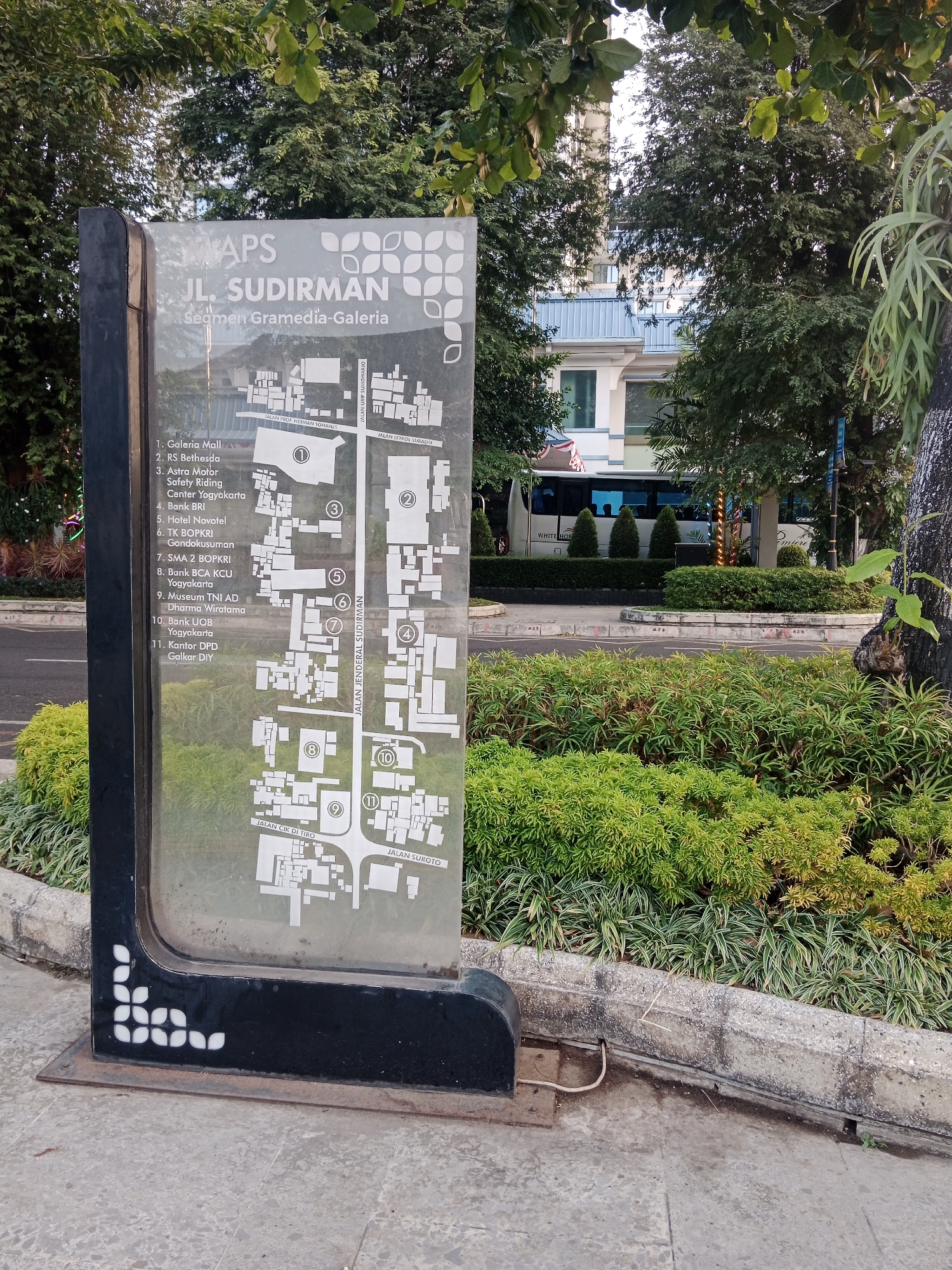 The image depicts an outdoor map sign located in front of a green landscaped area and a street, with a building and trees in the background. The map itself is titled "MAPS JL. SUDIRMAN" and specifies "Segmen Gramedia-Galeria". A numbered list of locations is visible on the left side of the sign, including "1. Galeria Mall", "2. RS Bethesda", and "3. Astra Motor Safety Riding Center Yogyakarta". The map illustration shows a layout of buildings and roads with corresponding numbers. A white bus with "WHITE HORSE" written on it is parked on the street behind the landscaping. The overall environment suggests a public urban setting in Yogyakarta, Indonesia.