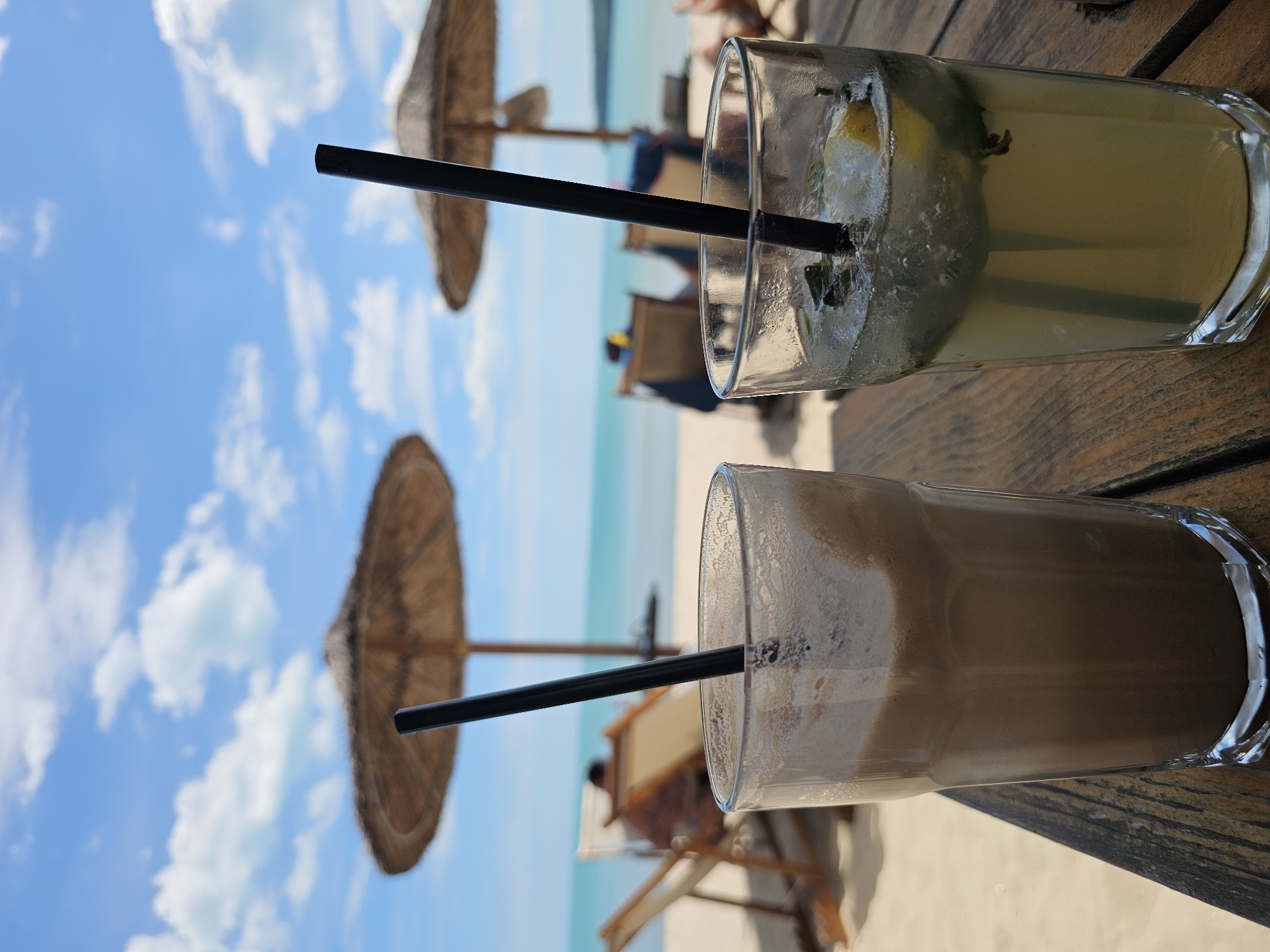 The image depicts a beach scene in Unknown, Unknown, with two drinks placed on a wooden table in the foreground. The drink in the top right of the frame is a light-colored liquid with ice cubes, garnished with lemon slices and mint leaves, and a black straw. The other drink, in the lower right, is a dark-colored liquid, likely coffee, with a frothy layer on top and a black straw. In the background, there is a beach with two straw umbrellas, beach chairs, and people seated under the umbrellas. The horizon shows the sea and sky. The sky is blue with scattered white clouds. The image is oriented horizontally, giving a wider view of the beach setting.