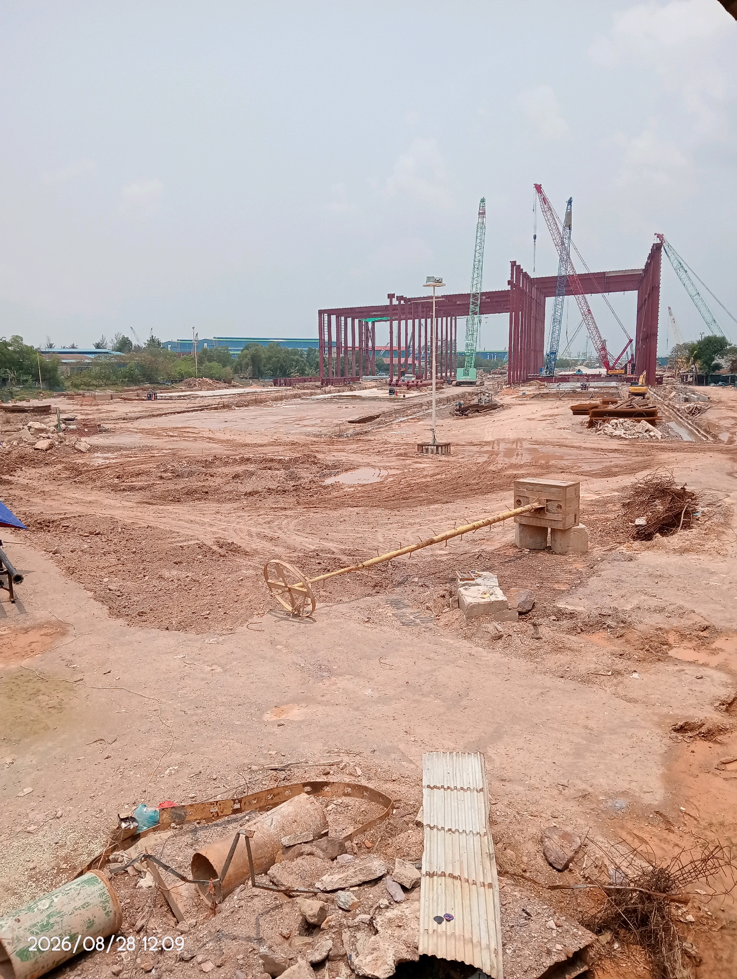 The image depicts a construction site in Batam, Indonesia. In the foreground, a yellow circular metal object on a pole is partially visible, resting on the muddy ground. Scattered debris, including pipes, concrete, and rebar, litters the area. In the mid-ground, a large-scale construction project is underway, with a massive red steel structure dominating the scene. Several large cranes, including green and red ones, are erected around the structure, suggesting active construction. The environment is characterized by uneven, dirt-covered terrain with puddles of water, indicating recent rain or poor drainage. In the background, industrial buildings and lush green trees are visible under a hazy, overcast sky. The timestamp "2026/08/28 12:09" is overlaid on the bottom left corner of the image, indicating the date and time the photograph was taken.