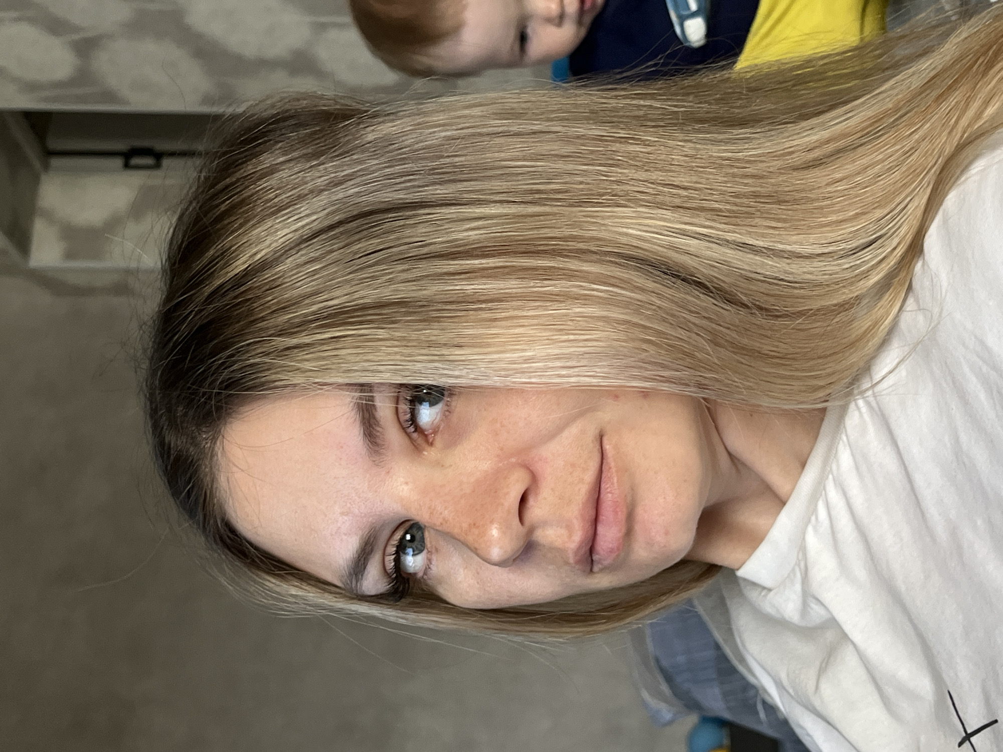 A woman with blonde hair is facing the camera. Part of a child's face is visible in the upper part of the image. The woman is wearing a white t-shirt. The background is a muted, neutral color. The image was taken indoors, in a room. There is no evidence to suggest this image was taken in Chaikovsky, Russia.