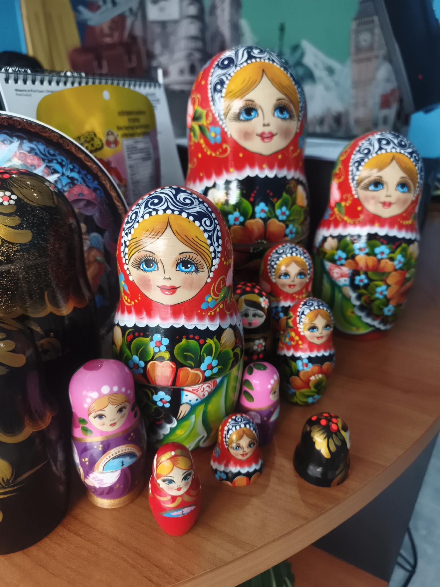 The image shows a collection of Russian nesting dolls (matryoshka dolls) of varying sizes arranged on a wooden surface. The dolls are painted with traditional folk designs, featuring female faces with blue eyes, red cheeks, and headscarves. The color palettes include red, black, green, gold, and pink. In the background, partially obscured, are a calendar, a package of snacks, and an image of what appears to be the Big Ben clock tower in London. The environment suggests an interior setting, possibly a home or souvenir shop. There is no evidence to confirm the location is in Baan Mae Nam, Thailand.