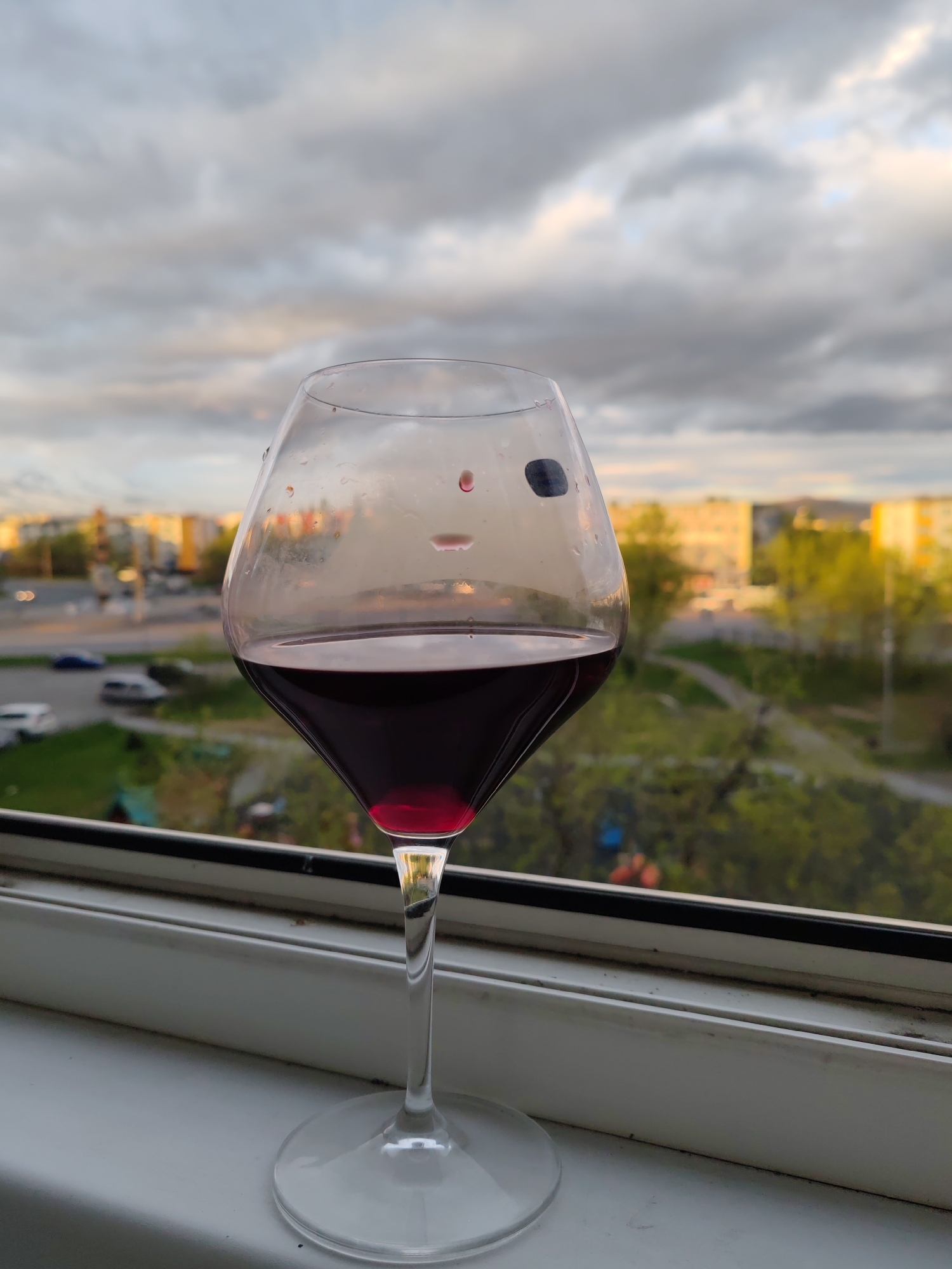 A glass of red wine sits on a windowsill with a cityscape in the background. The wine is dark red, filling the bottom half of the glass. The glass itself has condensation on it. The windowsill is white and appears to be made of plastic. Outside the window, buildings, trees, and a road can be seen. The sky is cloudy and appears to be dusk or dawn, with hints of orange and pink in the clouds. The scene is likely a residential area in the city of Petropavlovsk-Kamchatsky, Russia, as suggested by the building architecture and the overall urban landscape. There are no people or animals visible in the image. The lighting suggests it's either early morning or late evening.