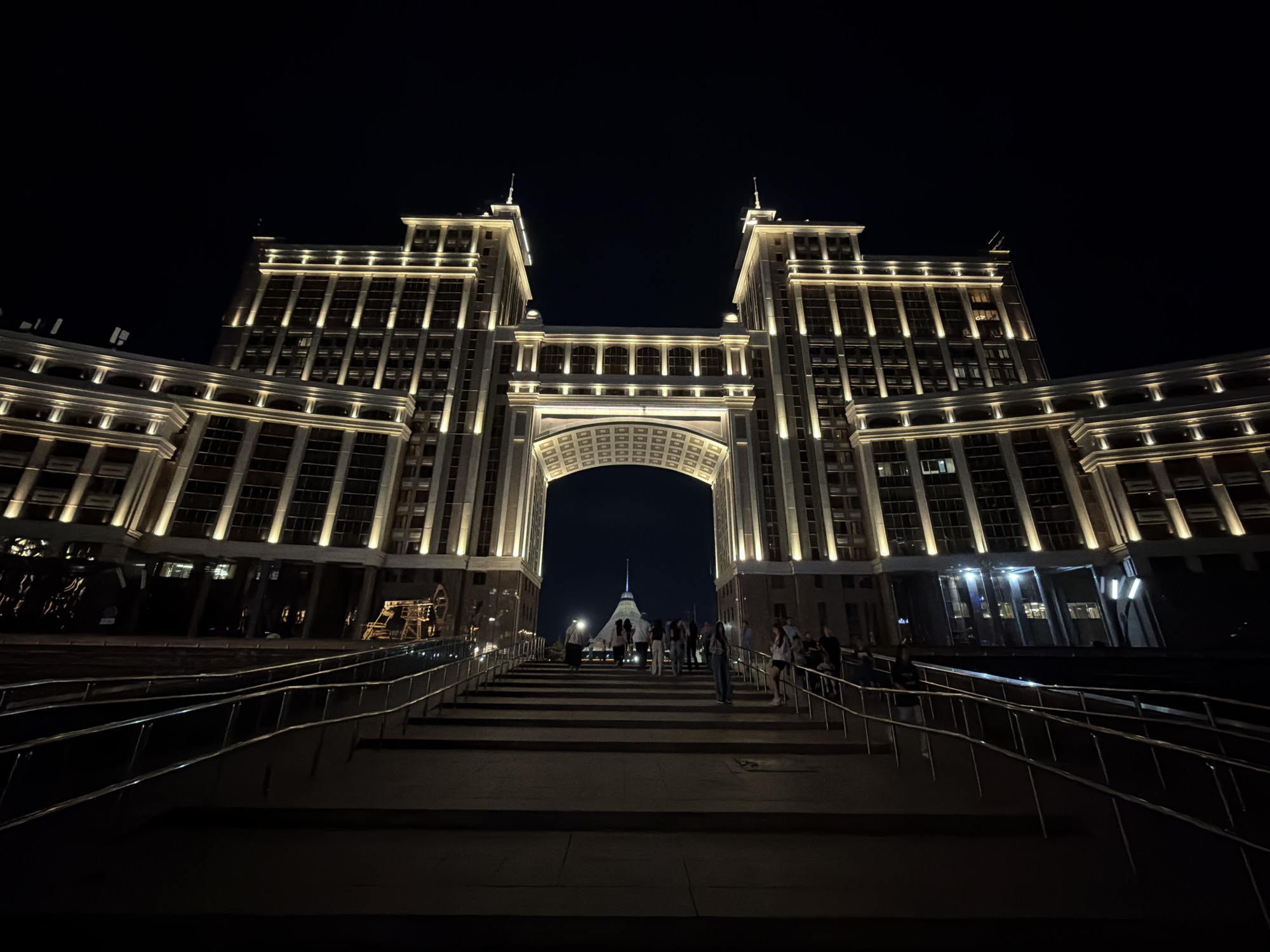 An exterior, low-angle, night shot captures the "Bayterek" monument in Astana, Kazakhstan, framed by an archway of a large, illuminated building. The building, constructed of beige stone, features multiple stories with rows of windows, and is lit by warm yellow lights along its facade, creating a dramatic and imposing presence. Two identical towers rise from the sides of the building, topped with pointed spires. The archway, a significant architectural element, is intricately designed with a patterned ceiling.
In the foreground, a wide, dark staircase leads up towards the archway, flanked by metal railings. Several people are visible on the stairs and at the top, appearing to be tourists or passersby, some in casual attire. Through the archway, the distinctive conical shape of the Bayterek monument is clearly visible, illuminated and standing out against the dark night sky. The overall scene evokes a sense of grandeur and urban nighttime beauty.