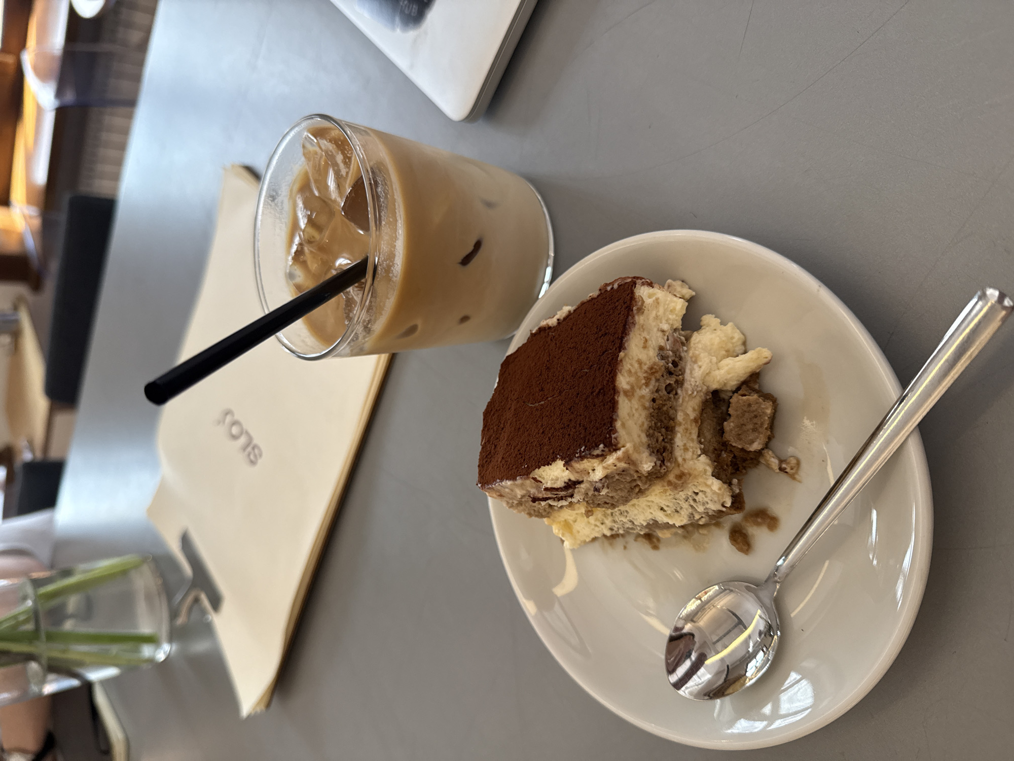 The image shows a table setting in what appears to be a cafe. The table surface is grey. A glass of iced coffee with a black straw sits on the left side, near a cream-colored paper bag. A slice of tiramisu is placed on a white plate to the right, accompanied by a silver spoon. Based on the "Sons" branding visible on the bag, the location is likely Sons Belgrade, a dessert shop in Belgrade, Serbia. No people are visible in the frame, though a vase with green stems is partially visible in the lower-left, suggesting someone is seated at the table. The environment appears to be indoors, given the lighting and table setting.