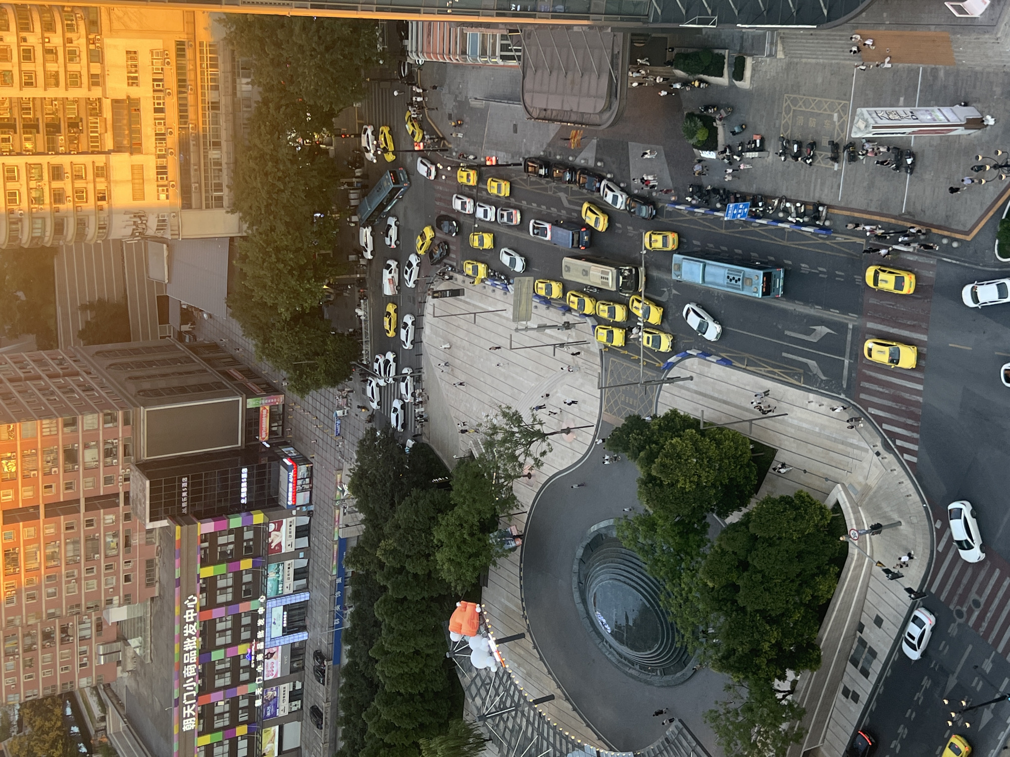 The aerial view captures a busy urban scene in Yuzhong District, China, during daylight. The composition is dominated by modern multi-story buildings with diverse architectural styles. One prominent building on the left features a colorful facade with numerous windows and commercial signage, including "朝天门小商品批发中心" (Chaotianmen Small Commodity Wholesale Center). Adjacent buildings display a mix of residential and commercial units, with some bathed in the warm glow of sunlight.
The ground level is a network of roads and pedestrian areas. Numerous vehicles, including yellow taxis, white cars, and a blue bus, are navigating the streets, suggesting moderate traffic congestion. Several people are visible walking on sidewalks and crossing streets. A large circular fountain with water features is a focal point in a paved plaza area, surrounded by trees and pedestrian walkways. In the plaza, there are large inflatable structures, one appearing to be orange and white. Further down the street, a row of parked motorcycles lines a pedestrian walkway. The scene is characteristic of a vibrant commercial and residential district in a Chinese city.