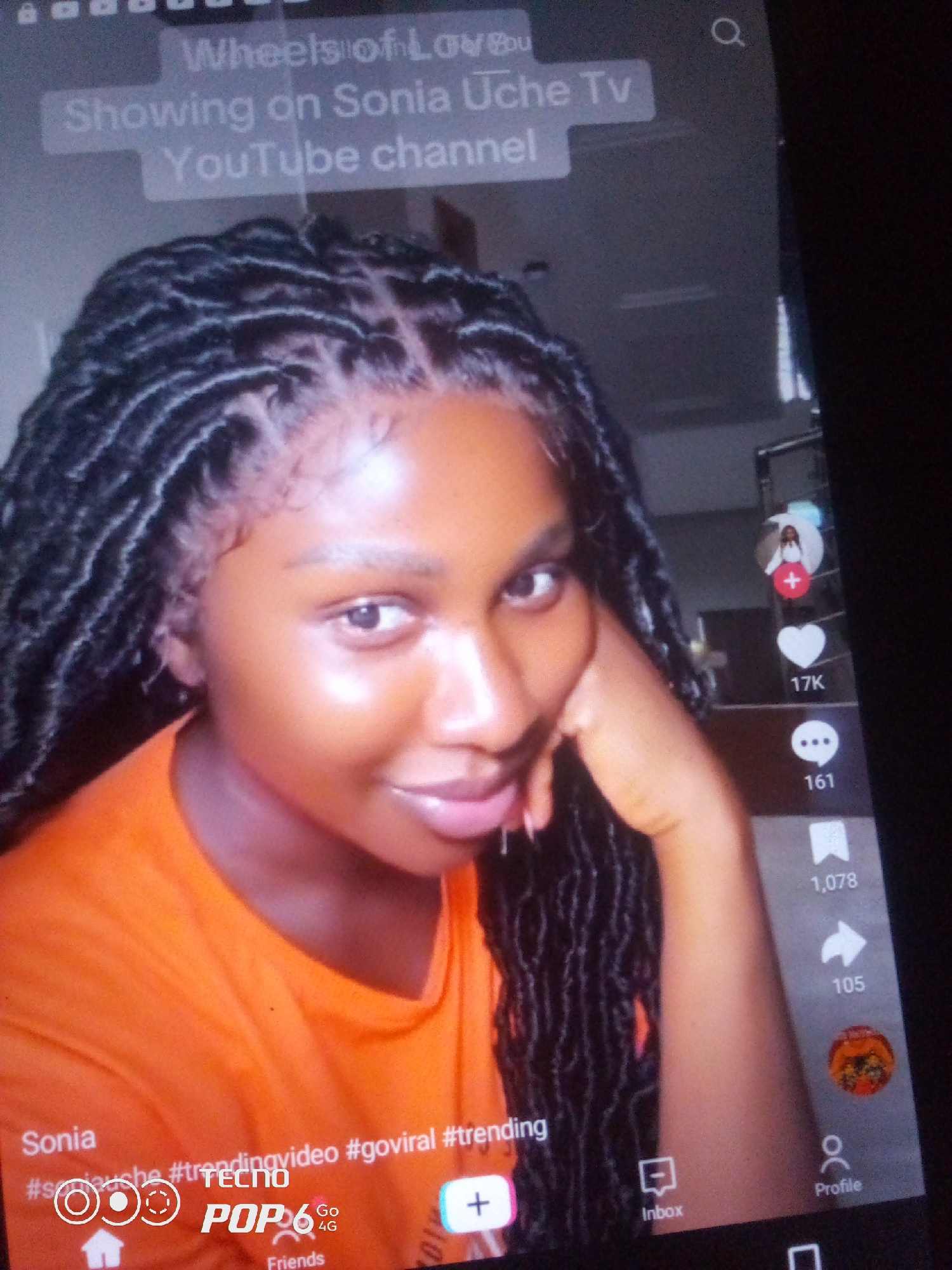 The image shows a close-up, slightly blurry, vertical photograph of a young Black woman with dark, braided hair, wearing an orange t-shirt. She is looking at the camera with a slight smile. Text overlays are visible on the screen, including "Wheels of Love," "Showing on Sonia Uche Tv," and "YouTube channel." On the right side of the screen, there are icons and numbers, suggesting a social media interface. These include a profile icon, a plus sign, a heart icon with "17K" next to it, a speech bubble icon with "161" next to it, and a bookmark icon with "1,078" next to it. At the bottom of the screen, the text "Sonia" is visible, followed by hashtags such as "#soniauche," "#trendingvideo," and "#goviral." There are also icons representing a house, a speech bubble, and a profile. The overall scene suggests a mobile phone screen displaying a video or social media post. There is no discernible information in the image to indicate the location within Howayi, Nigeria.