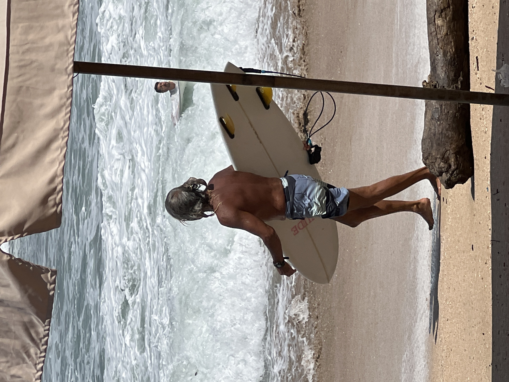 This image depicts a man with gray hair, bare-chested and wearing patterned swim trunks, walking on a sandy beach in Kuta, Indonesia. He is carrying a white surfboard under his right arm. The ocean is behind him, with waves breaking and foam on the water. In the background, another person is visible in the water, partially submerged and facing towards the shore. An umbrella with a tan fabric canopy is positioned in the foreground, casting a shadow on the sand. A weathered log lies on the beach in the lower right portion of the frame.