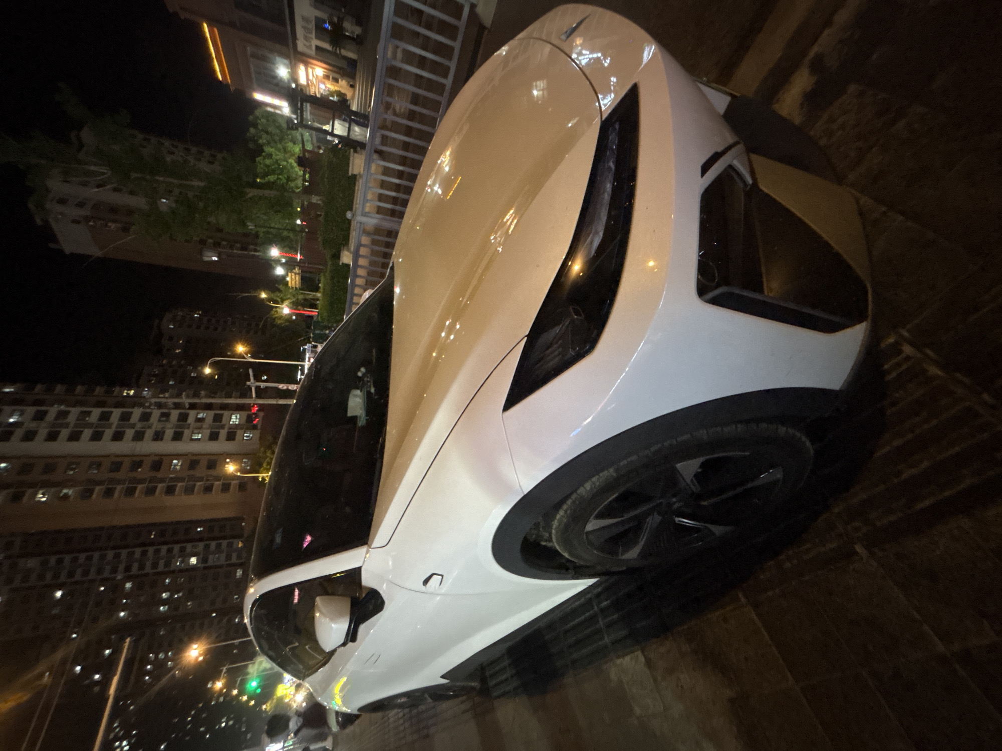 A white, modern-looking car is parked on a tiled sidewalk at night in Suzhou Industrial Park, China. The car's headlights are on, illuminating the street and the front of the vehicle. In the background, tall apartment buildings are visible, their windows lit up, suggesting a residential area. A metal fence and some greenery are also present, separating the sidewalk from what appears to be a street or public area. The streetlights and building lights create a somewhat illuminated, urban night scene.