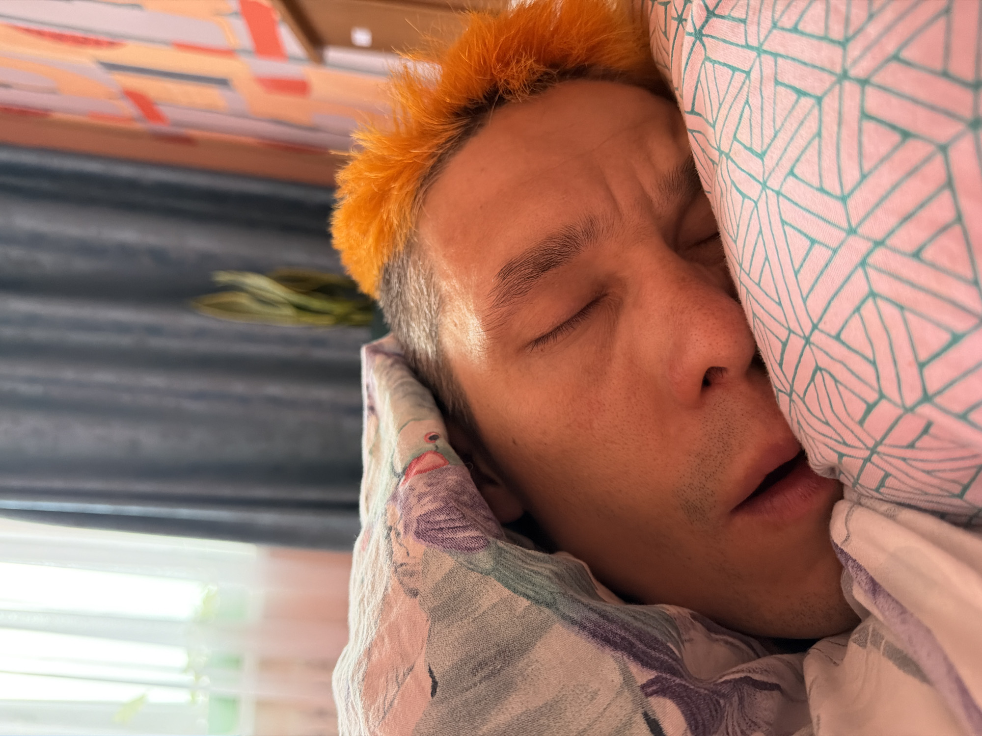 A close-up shot depicts a man sleeping in a bed. He has short, bright orange hair on top, transitioning to gray at the sides. His eyes are closed, and his mouth is slightly open. He is lying against a pillow with a geometric pattern of pink and teal lines. A floral-patterned blanket is draped around him. The background reveals a partially visible window with green foliage visible outside, and a curtain. The location is uncertain; the city of Chaikovsky, Russia, cannot be confirmed based on the visual data provided.
