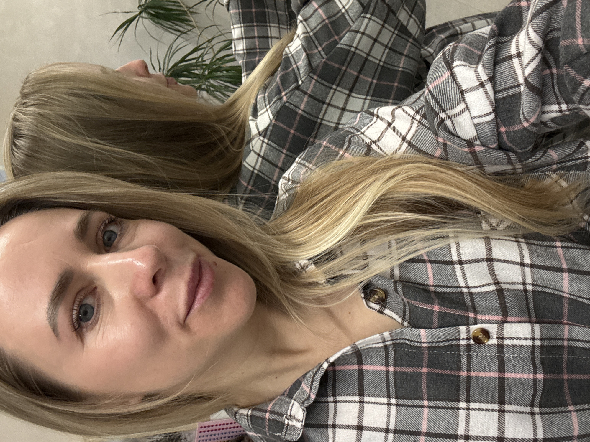 A close-up image features a woman with long, wavy blonde hair and blue eyes, looking directly at the camera with a neutral to slightly pleasant expression. She has a fair complexion.
She is wearing a long-sleeved plaid shirt with a pattern of grey, white, and pink lines. Brown buttons are visible on the shirt's placket and collar.
In the background, a distinct reflection of the same woman is visible, showing her in profile looking towards the right side of the frame, indicating she is standing before a mirror. Behind the reflection, a green leafy houseplant, possibly a type of palm or fern, is discernible, suggesting an indoor setting. The overall lighting is soft and even.
The image is presented with the woman's head tilted towards the right. There is no visible text in the media file. The context provided indicates the location is Chaikovsky, Russia.