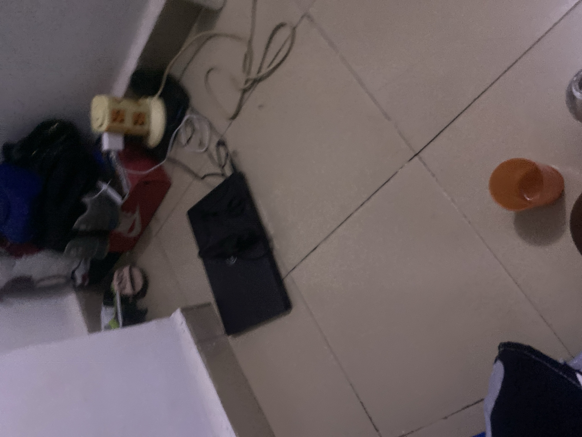 The image depicts a floor with beige tiles in a dimly lit room. In the upper left corner, a white wall meets the floor. The room appears to be a living space or bedroom, with several items scattered around. A black laptop is open and placed on the floor in the center of the frame. To the right of the laptop, a yellow cylindrical object with orange markings, possibly a fan or air freshener, is plugged into an outlet. Various cords are strewn across the floor around the electronic devices. In the background, there are shoes and bags. In the lower right corner, an orange cup is visible. The overall impression is one of a cluttered domestic environment. The specific location of Howayi, Nigeria, cannot be determined from the visual information alone.