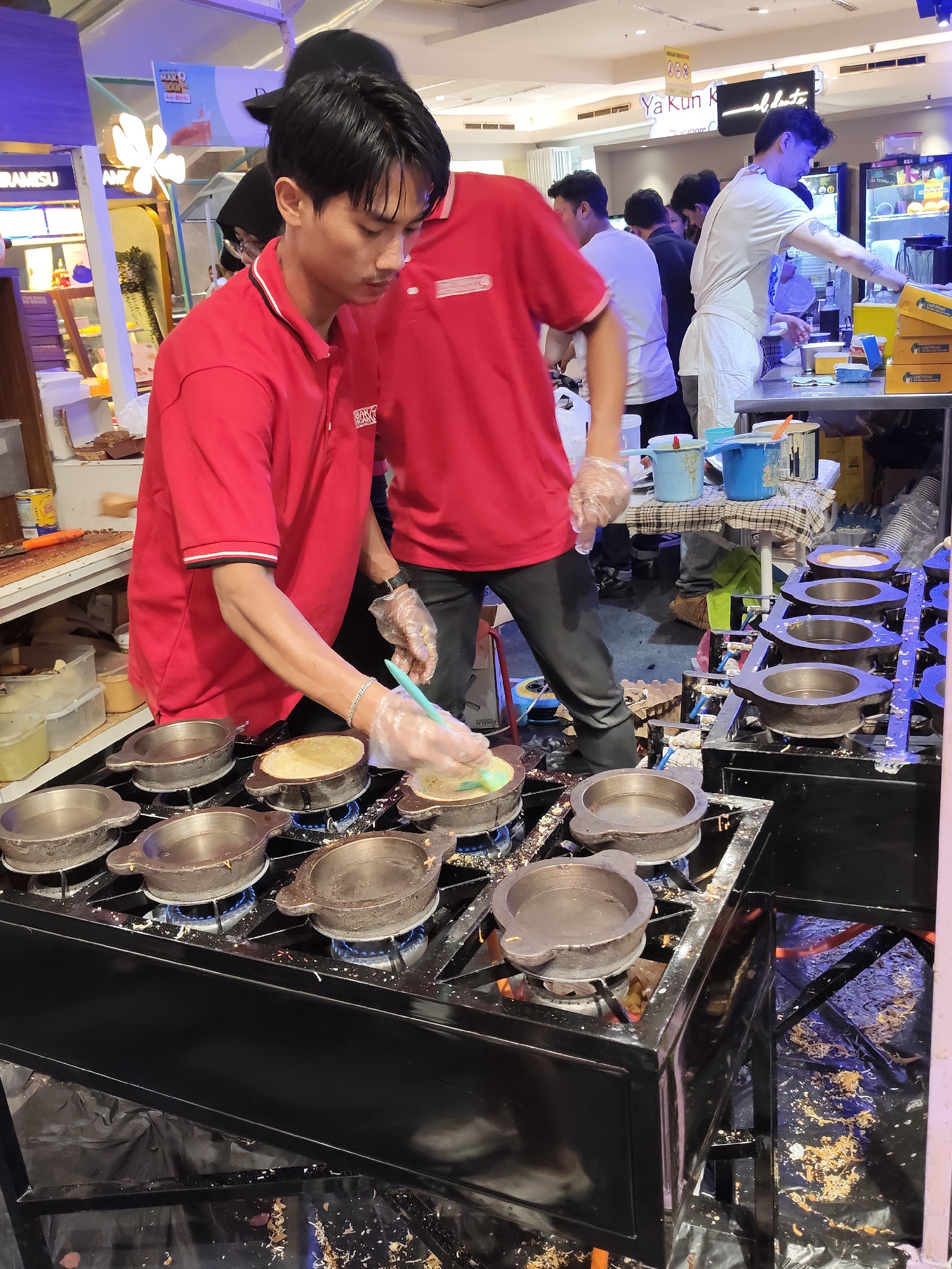In a well-lit indoor market or food court in the City of Medan, Indonesia, a street food vendor, identified by his red uniform, is actively preparing food. He is wearing clear plastic gloves and is in the process of cooking what appears to be a pancake-like dish in a specialized multi-mold griddle. The griddle, which has eight circular molds, is placed on top of a commercial gas stove. The vendor is holding a blue brush and seems to be spreading batter into one of the molds. Another person in a similar red uniform is visible behind him. In the background, other individuals are seen mingling, and various food stall displays are present, including signage that reads "Ya Kun Kopi". The area around the stove appears somewhat messy, with scattered food debris and packaging.