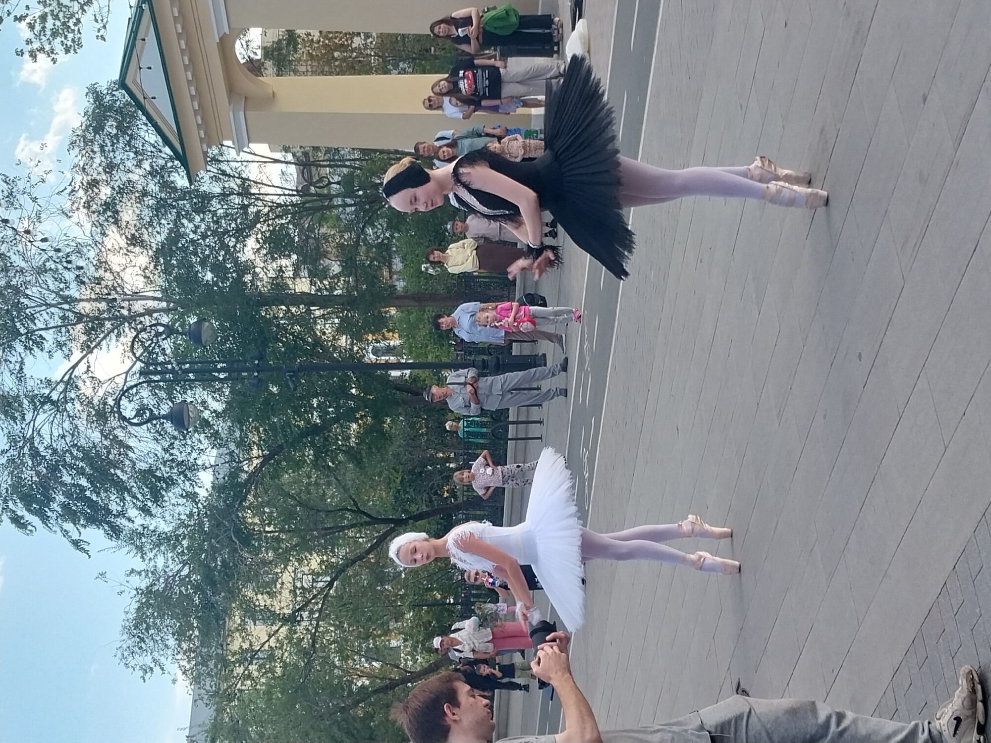 In Orenburg, Russia, two young ballerinas, one in a white tutu and the other in a black tutu, perform on a paved outdoor area. The white tutu ballerina is positioned slightly to the left, facing mostly forward, while the black tutu ballerina is to the right, facing slightly to the left. Both are wearing pointe shoes and have their hair styled. A man holding a camera is visible on the far left, seemingly filming the performance. A crowd of spectators, including adults and children, watches from the background, some standing and others sitting. Behind the performers and spectators, trees line the area, and a building with an archway is visible on the right. The sky above is a clear blue with some clouds. The overall scene depicts a public performance of ballet in a park-like setting within the city.