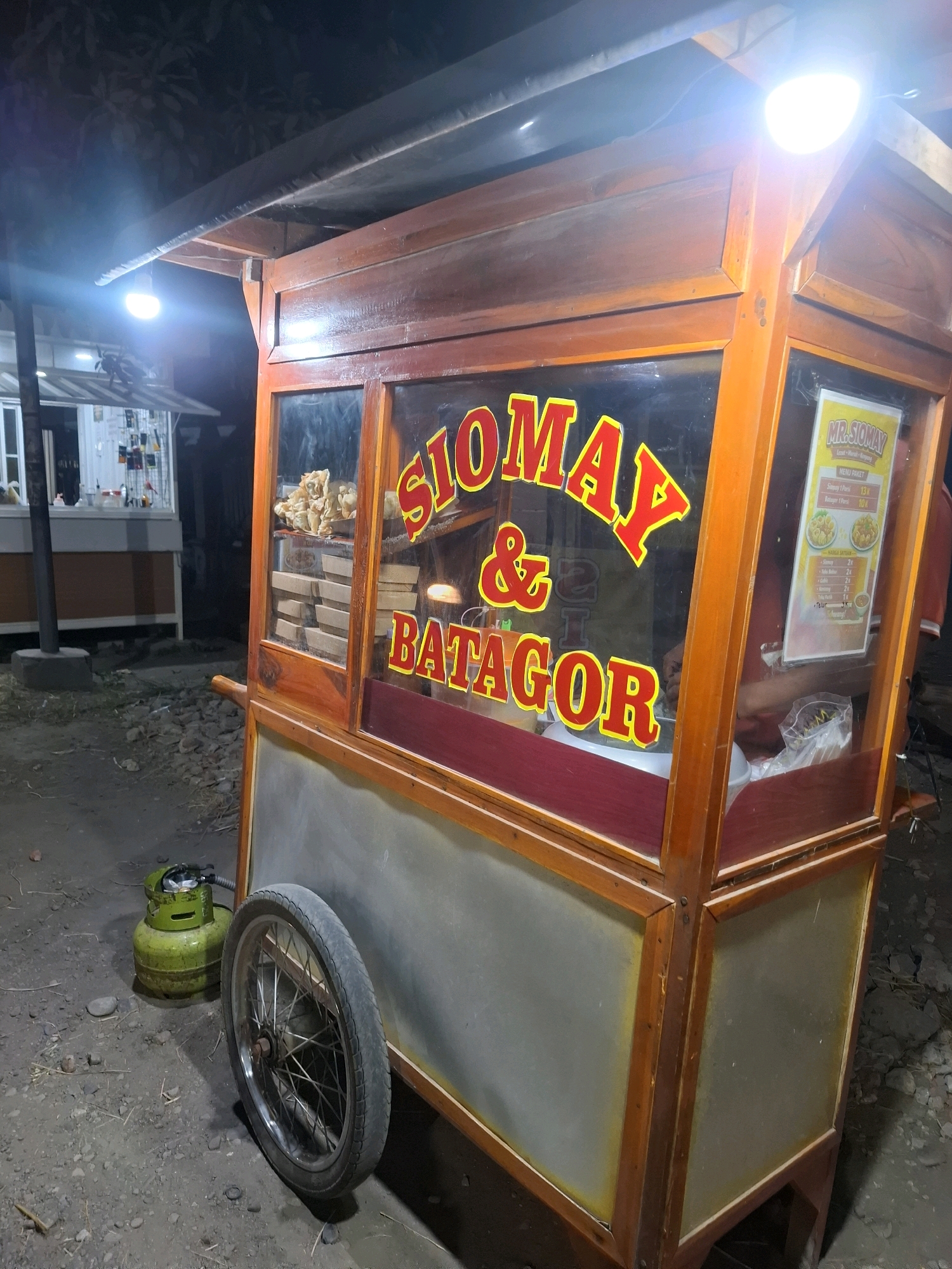 In Simo, Indonesia, a mobile food cart, likely serving "Siomay & Batagor," is illuminated at night by bright overhead lights. The cart is constructed of wood with glass windows displaying food items and a menu. A green propane tank sits on the gravelly ground to the left of the cart, next to a bicycle wheel. In the background, another food stall or structure is visible.
